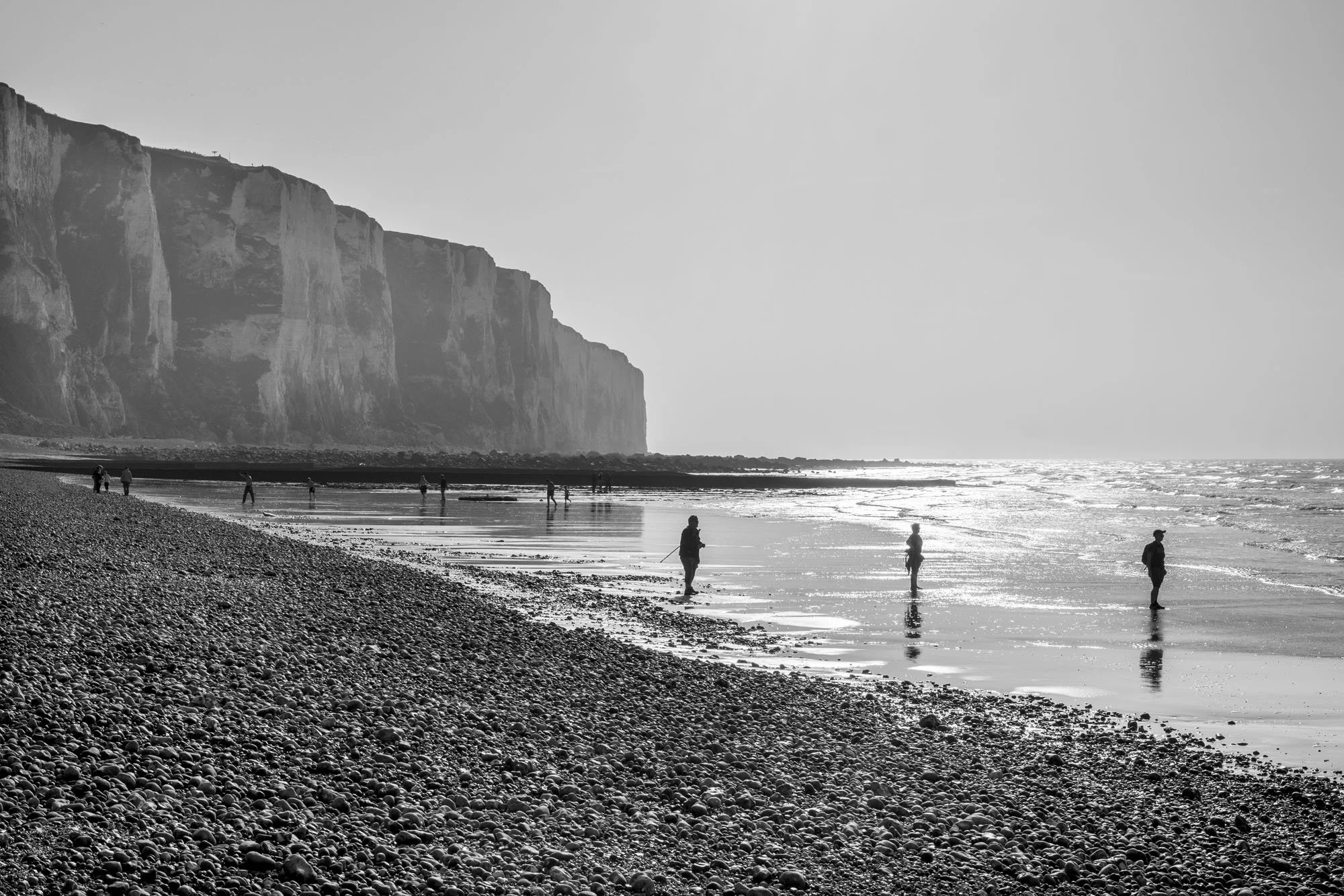  Black-and-white seaside photograph on the beach at Le Tréport, Normandy, three men’s silhouettes at the pebble beach, by Chris Perona. 