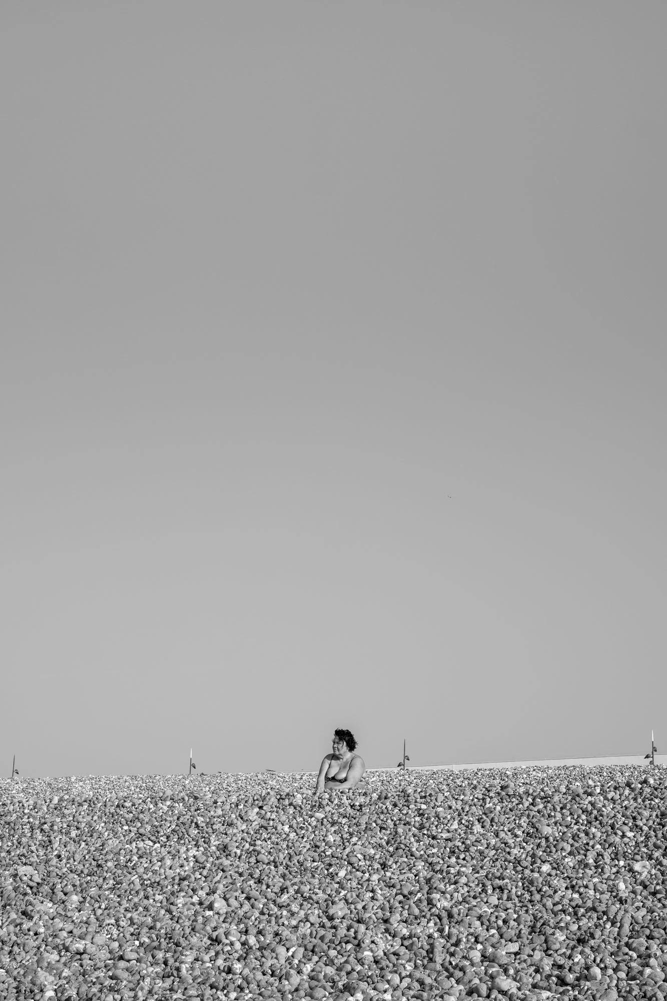 Black-and-white seaside photograph on the beach at Le Tréport, Normandy, a lonely woman seated hidden partially by the pebble dune,  by Chris Perona. 