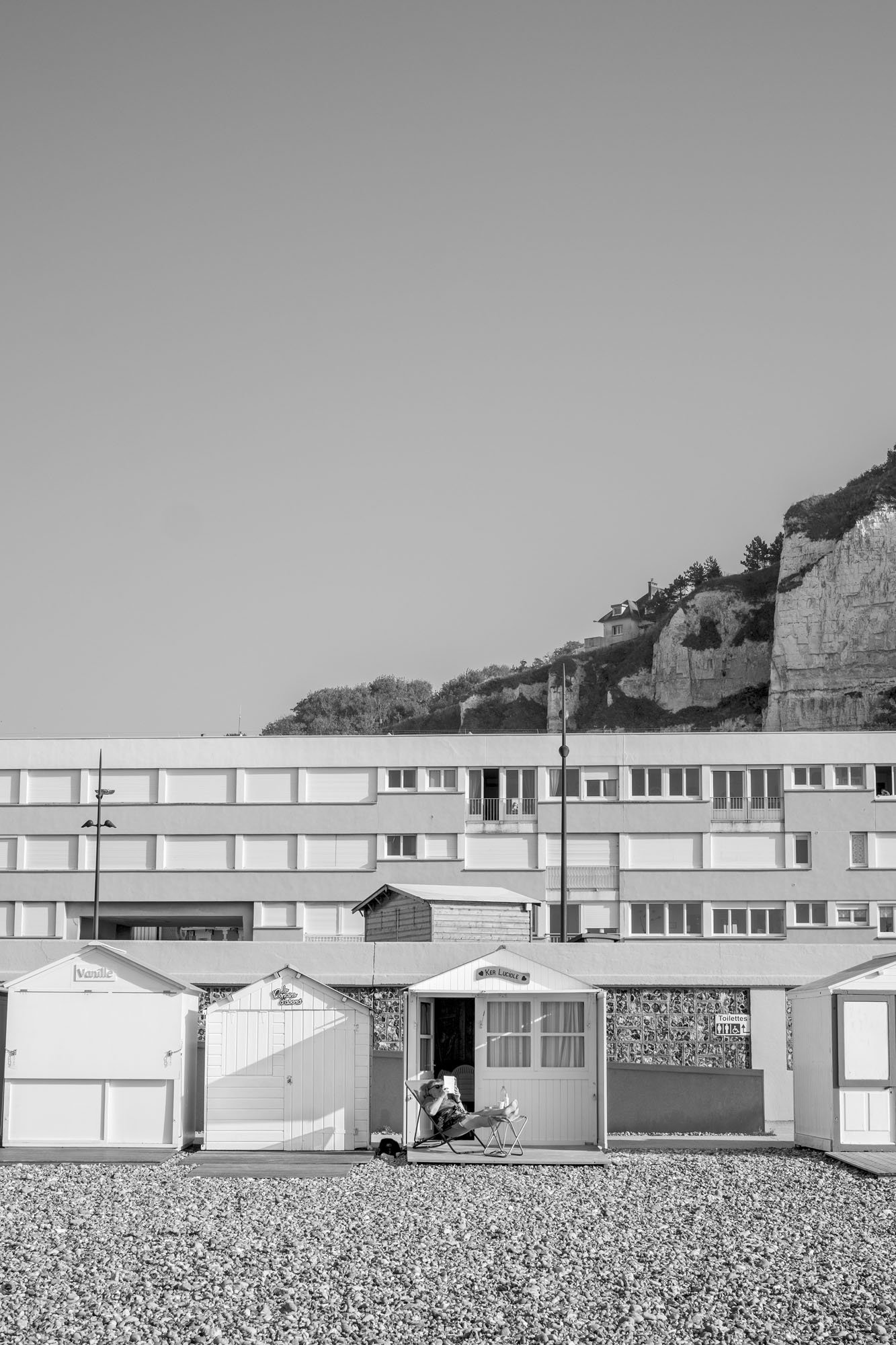  Black-and-white seaside photograph on the beach at Le Tréport, Normandy, a lonely person reading and sunbathing with the city and hill in the background, by Chris Perona. 