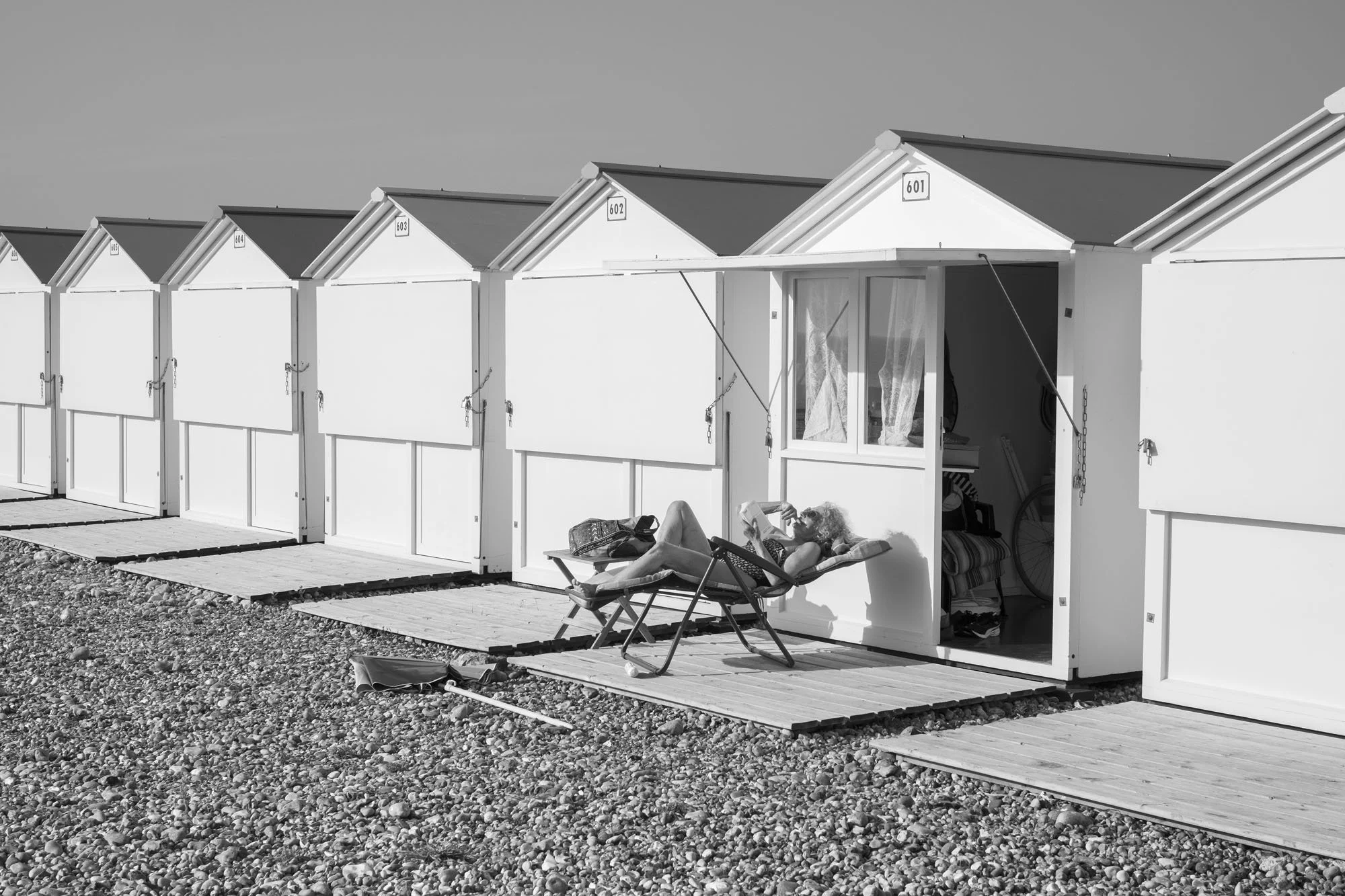  Black-and-white seaside photograph on the beach at Le Tréport, Normandy, an old lady sunbathing and reading by a row of beach huts, by Chris Perona. 