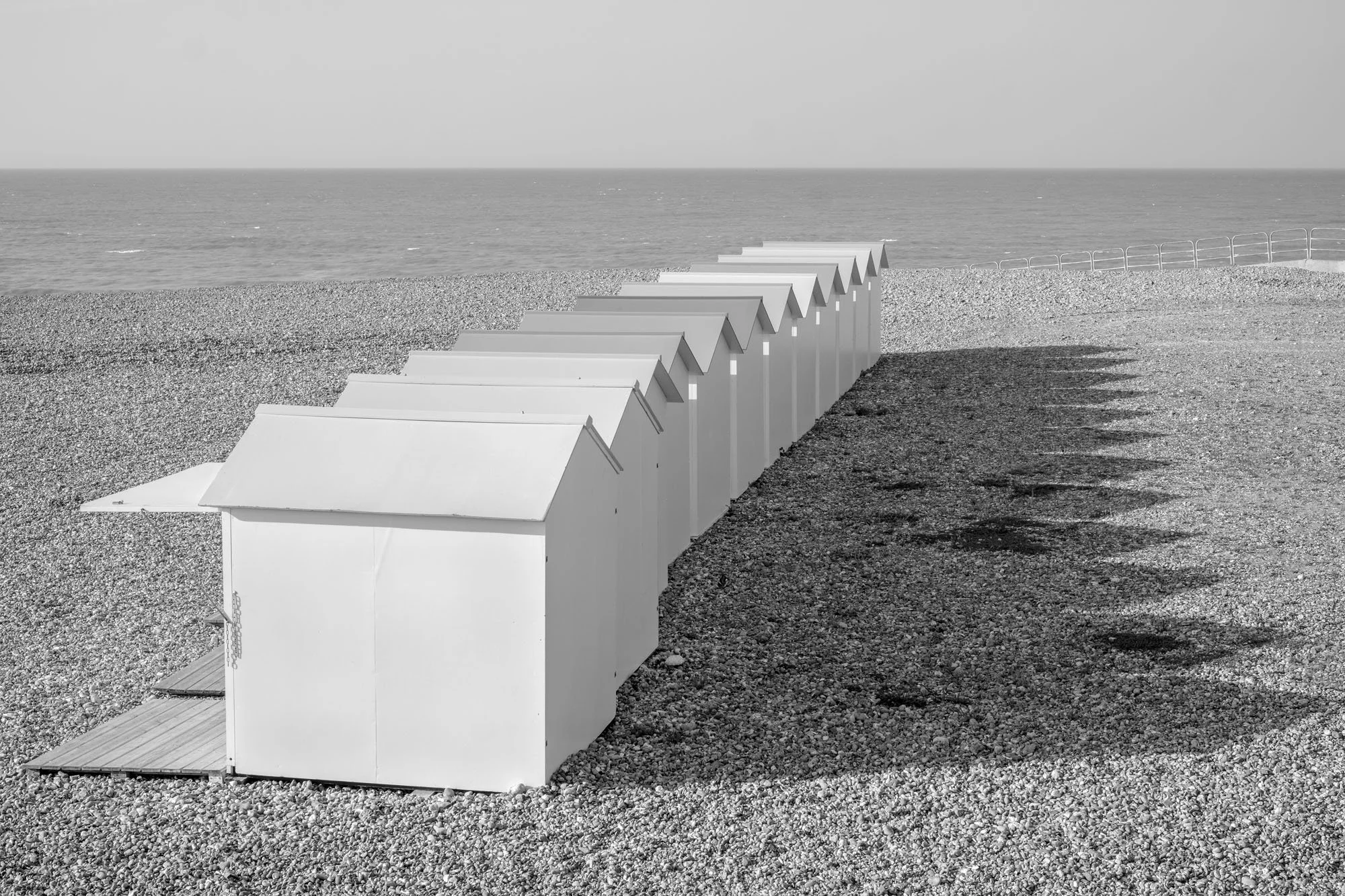  Black-and-white seaside photograph on the beach at Le Tréport, Normandy, a row of beach huts and their shade, by Chris Perona. 