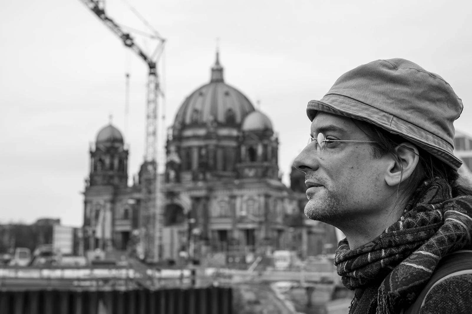  Man in a bucket hat and scarf in profile, with the Berlin Cathedral and a crane behind him. Candid street portrait by Chris Perona. 