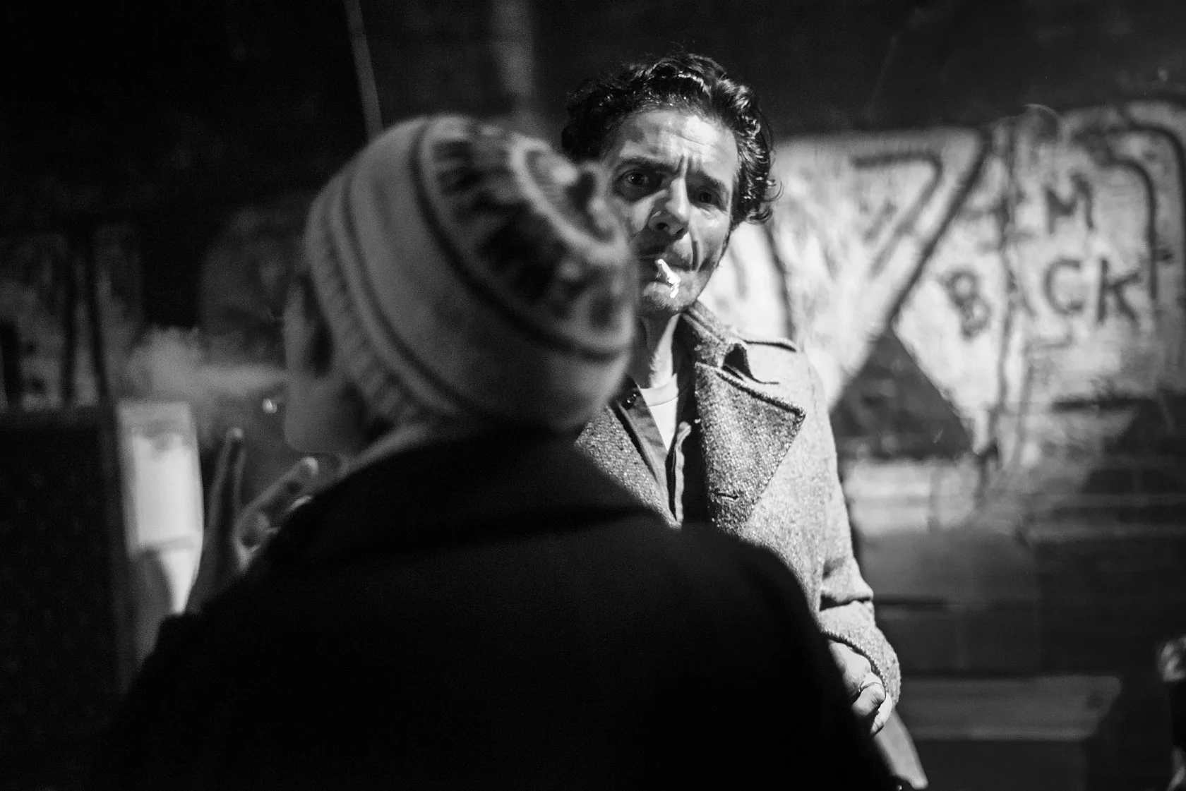  Man in a coat talking with a friend in a knit hat by a graffiti wall at night, Berlin. Candid street portrait by Chris Perona. 