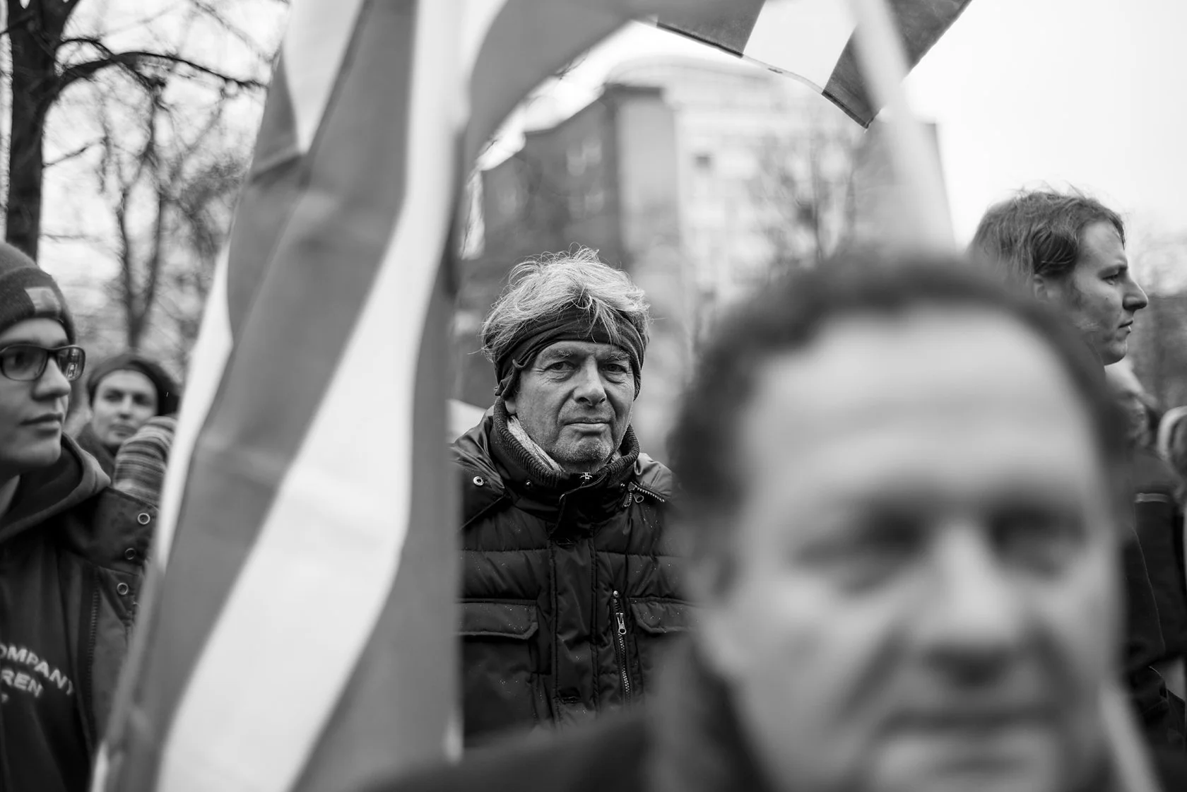  Men among flags at a demonstration, one face close in the foreground, Berlin. Candid street portrait by Chris Perona. 