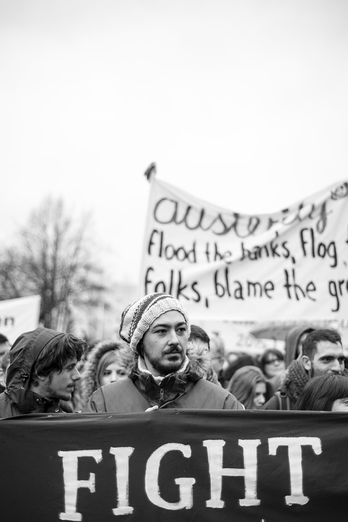  Man in a knit hat marching behind a "Fight" banner at a protest, Berlin. Candid street portrait by Chris Perona. 