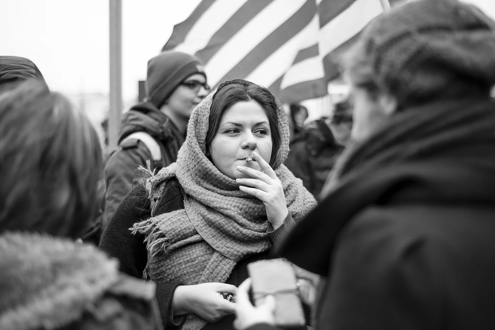  Young woman wrapped in a knitted scarf, smoking in a crowd beneath a flag, Berlin. Candid street portrait by Chris Perona. 