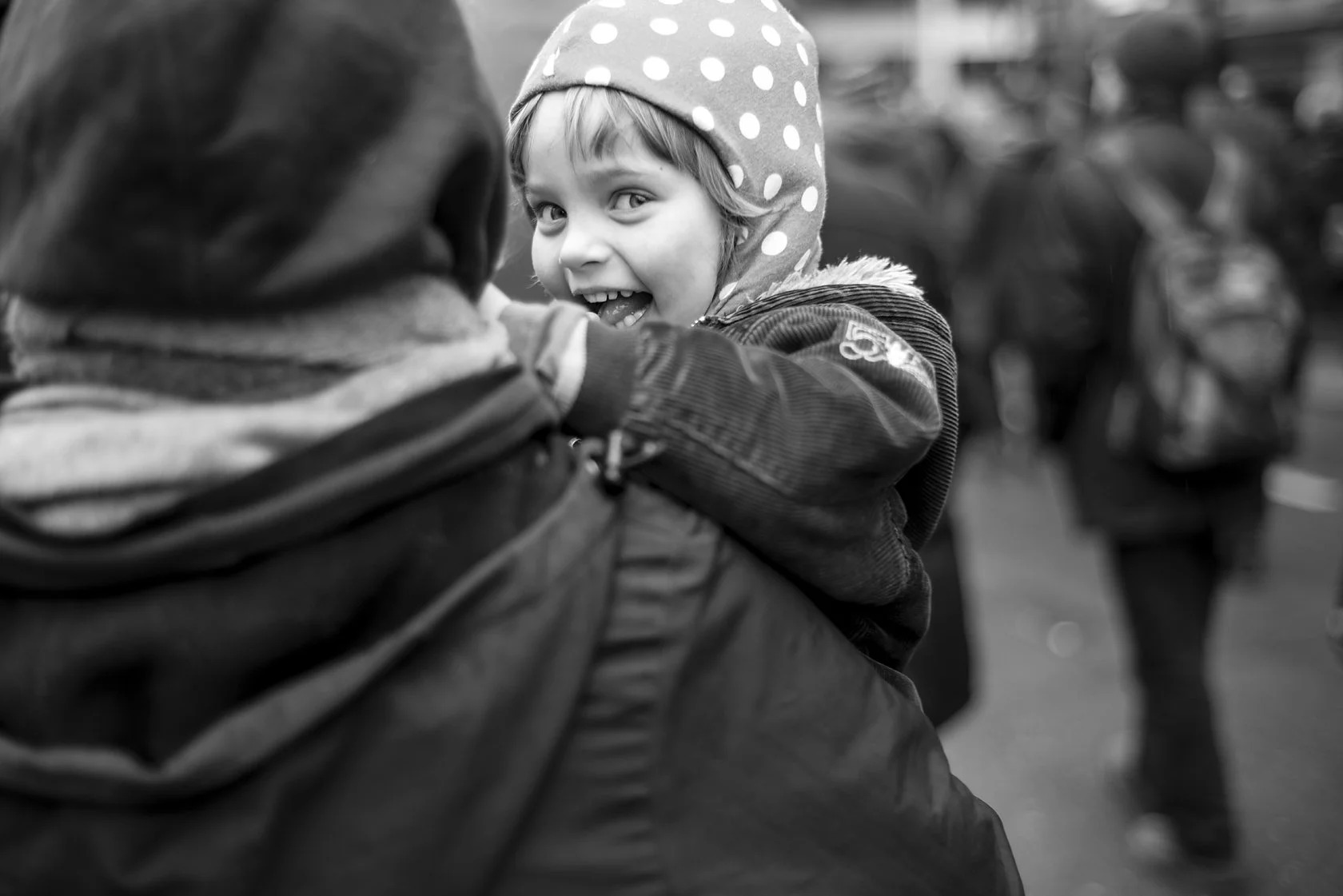  Laughing child in a polka-dot hat carried through a crowd, Berlin. Candid street portrait by Chris Perona. 