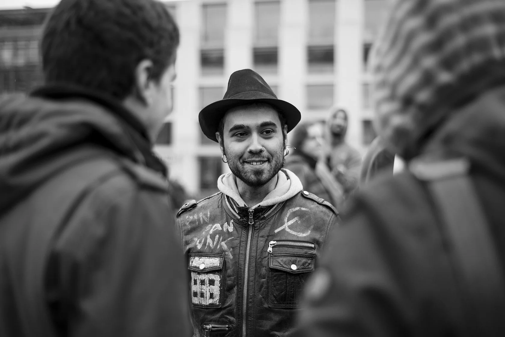  Smiling man in a hat and patched leather jacket in a crowd, Berlin. Candid street portrait by Chris Perona. 