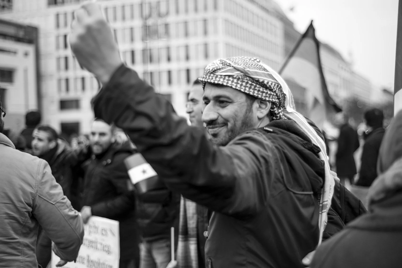  Smiling man in a checkered scarf raising his arm at a street demonstration, Berlin. Candid street portrait by Chris Perona. 