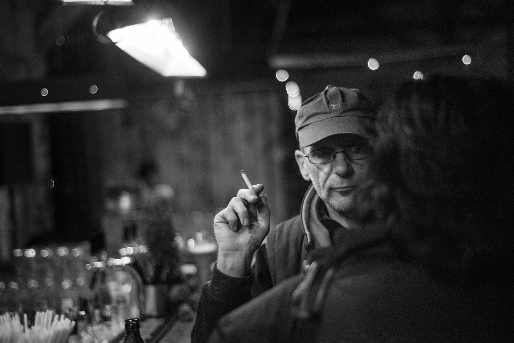  Older man in a cap smoking and talking in a dimly lit bar, Berlin. Candid street portrait by Chris Perona. 