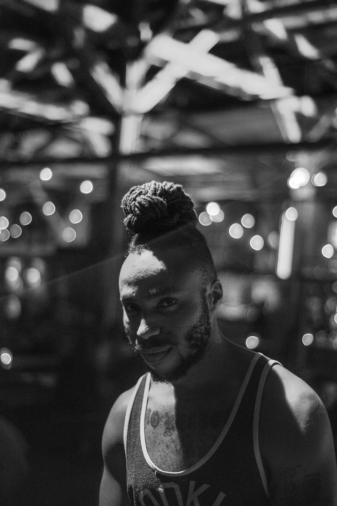  Man with tied-up braids and a tank top, smiling under string lights at night, Berlin. Candid street portrait by Chris Perona. 