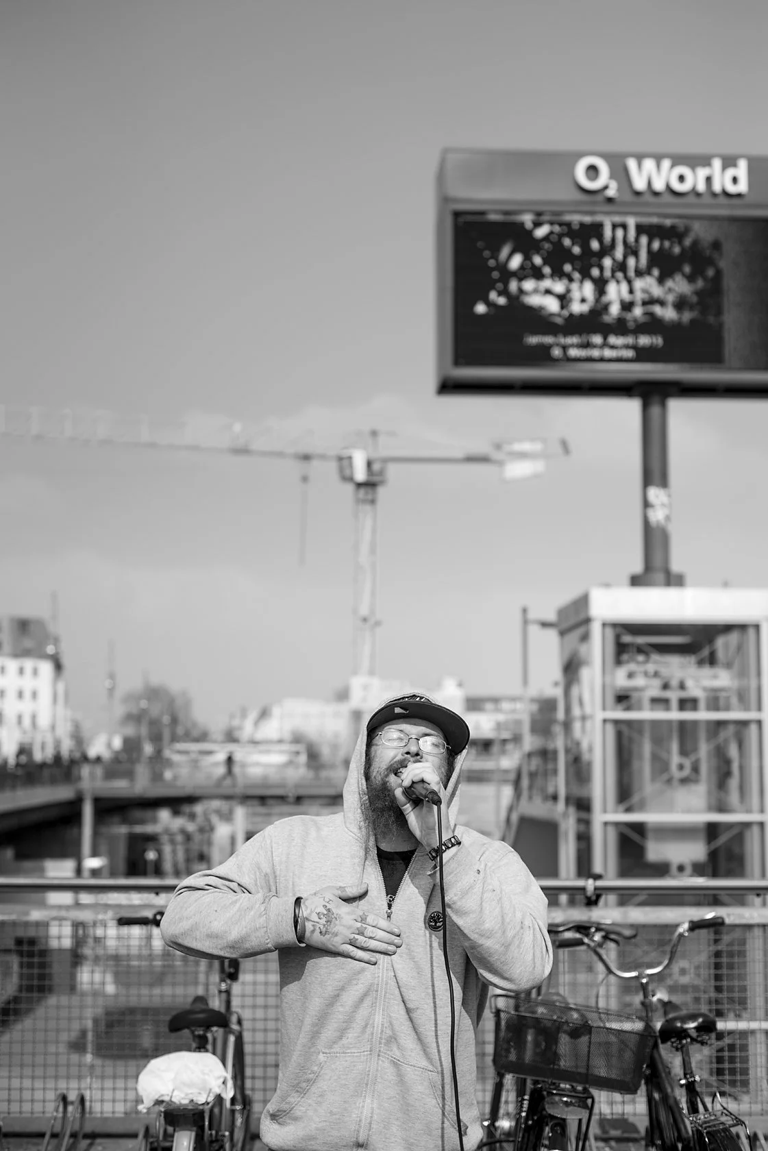  Street singer in a hoodie and cap singing into a microphone near the O2 World arena, Berlin. Candid street portrait by Chris Perona. 