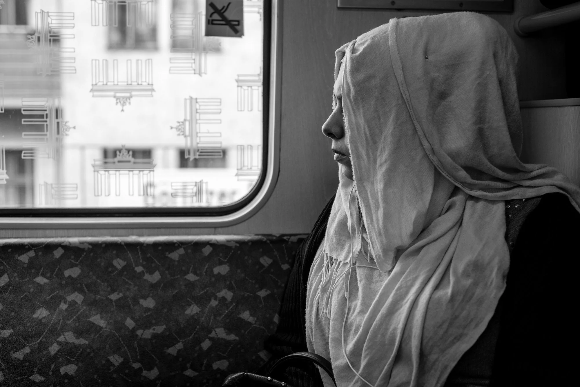  Woman with her head wrapped in a light scarf, seated by a train window in Berlin. Candid street portrait by Chris Perona. 