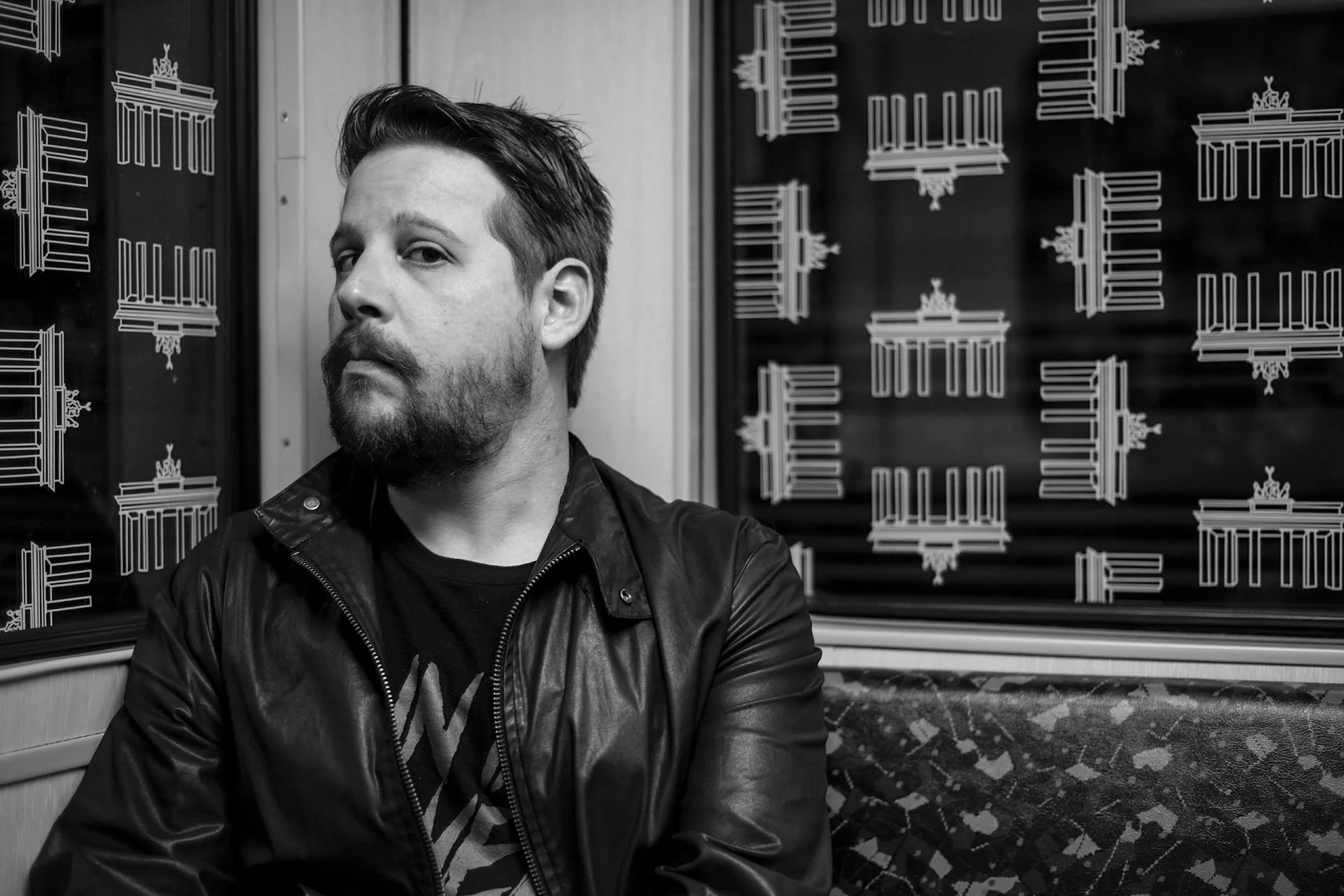  Bearded man in a leather jacket on a Berlin U-Bahn, Brandenburg Gate pattern on the seats behind him. Candid street portrait by Chris Perona. 