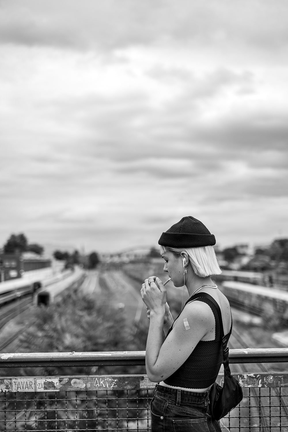  Young woman in a black beanie lighting a cigarette on a bridge above railway tracks, Berlin. Candid street portrait by Chris Perona. 