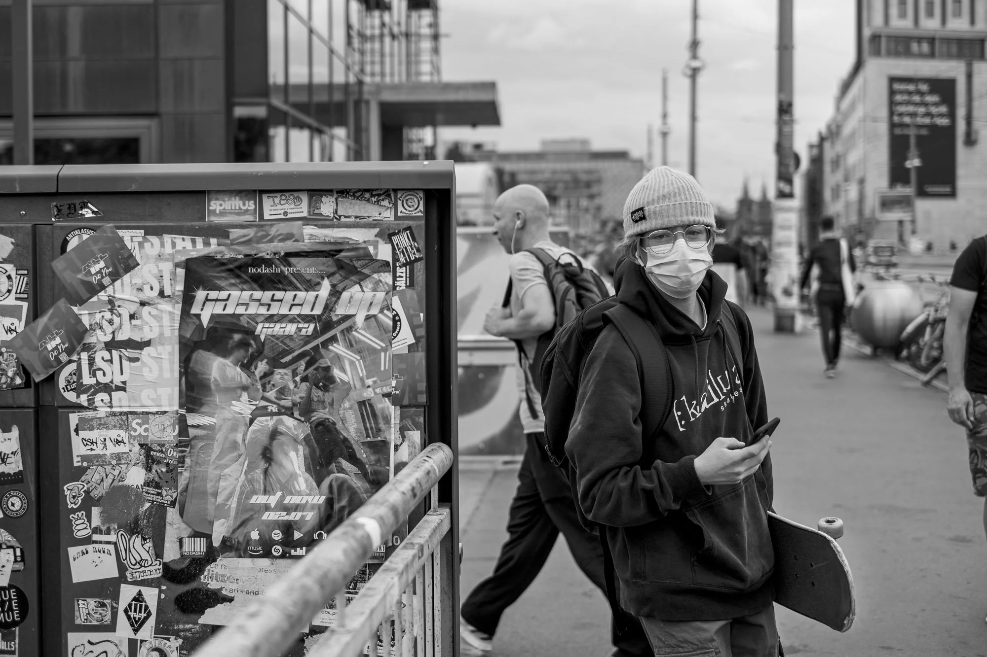  Young man in a face mask carrying a skateboard past a sticker-covered box, Berlin. Candid street portrait by Chris Perona. 