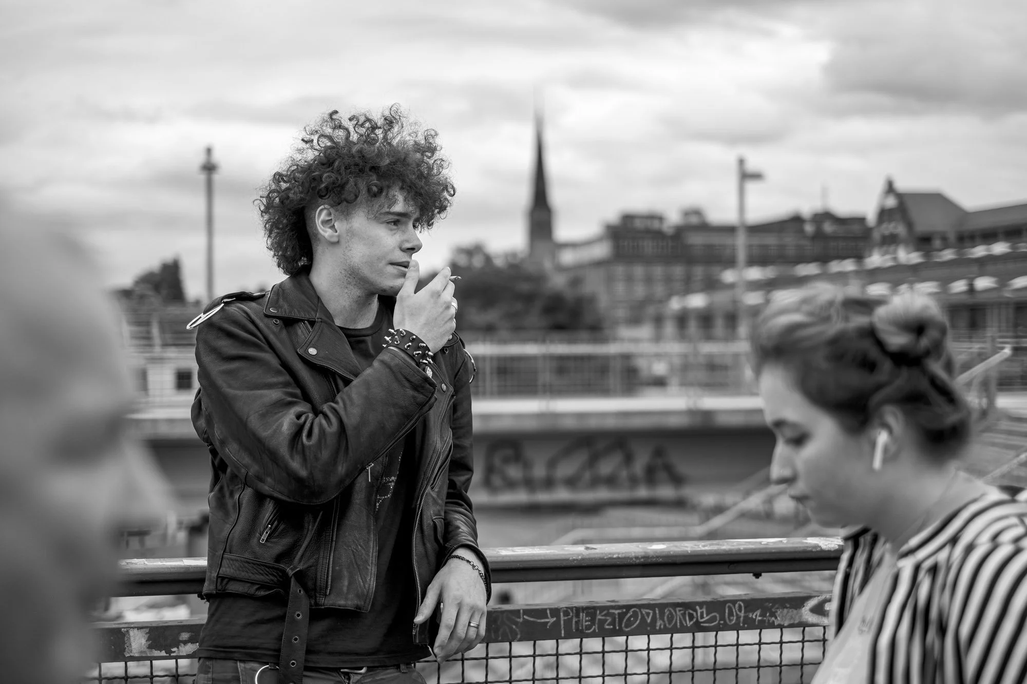  Young man with curly hair smoking by the river, a church spire behind him, Berlin. Candid street portrait by Chris Perona. 
