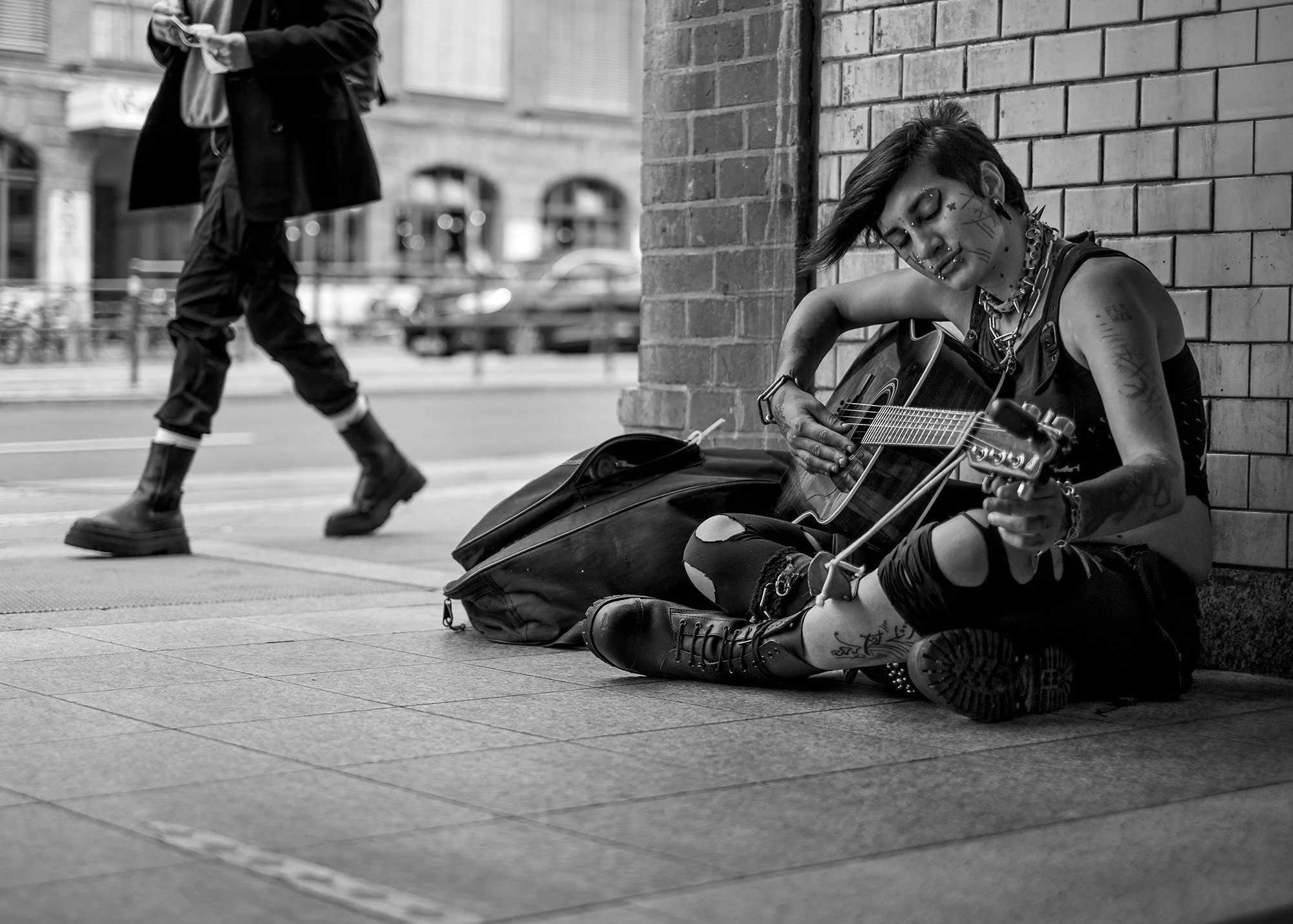  Street musician playing guitar, seated against a tiled wall as a passer-by walks on, Berlin. Candid street portrait by Chris Perona. 