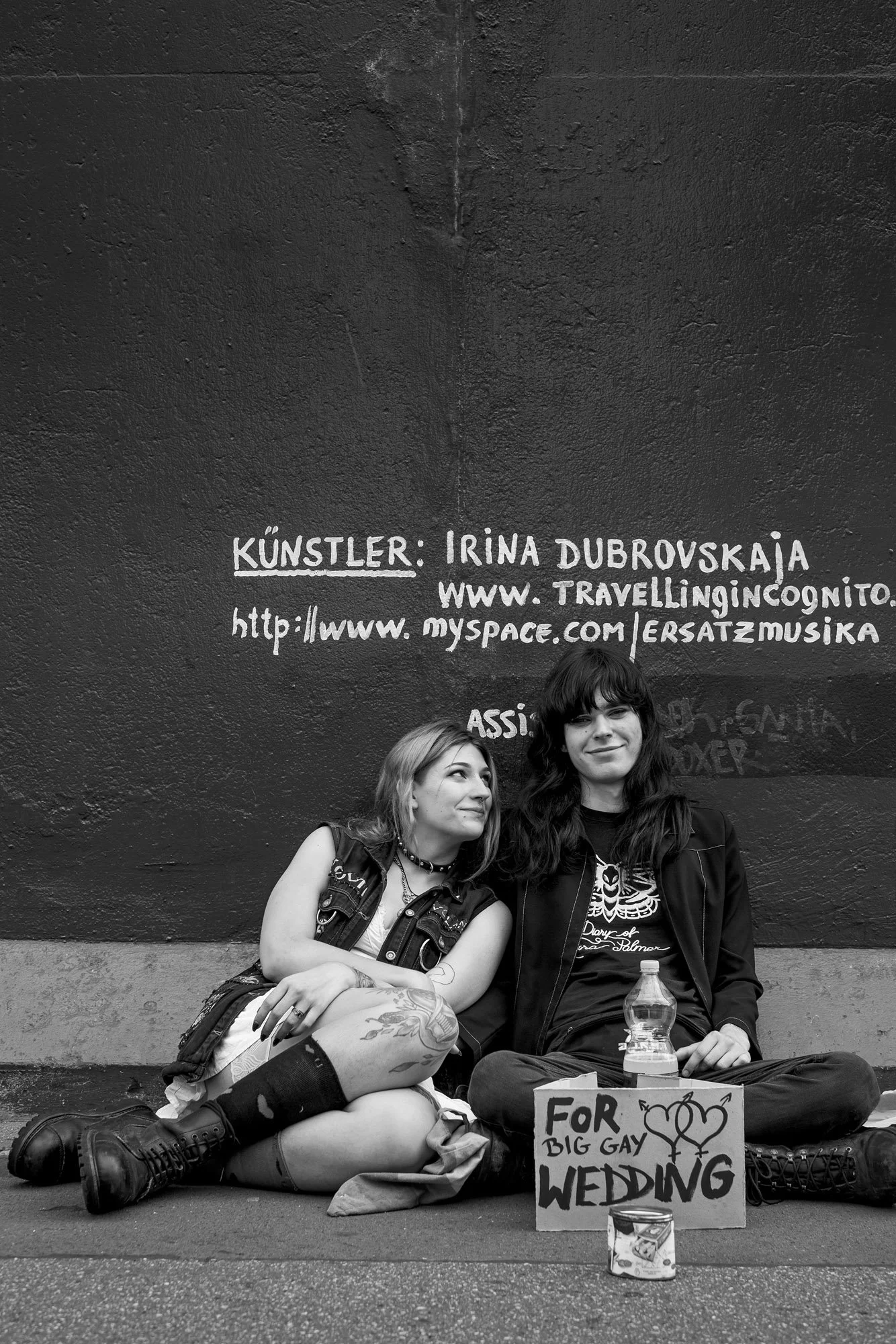  Two young women sitting on the pavement with a handwritten sign about their wedding, Berlin. Candid street portrait by Chris Perona. 