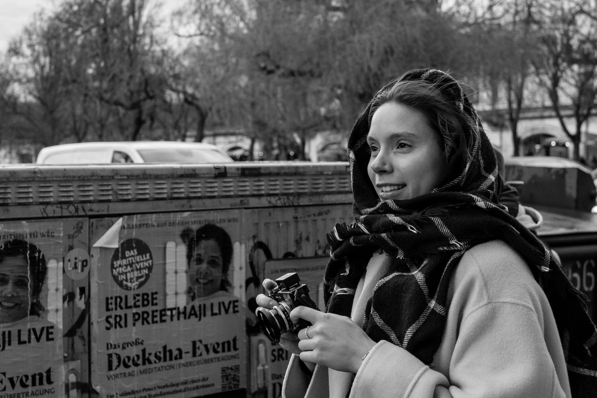  Smiling young woman in a plaid scarf holding a vintage camera beside a poster wall, Berlin. Candid street portrait by Chris Perona. 