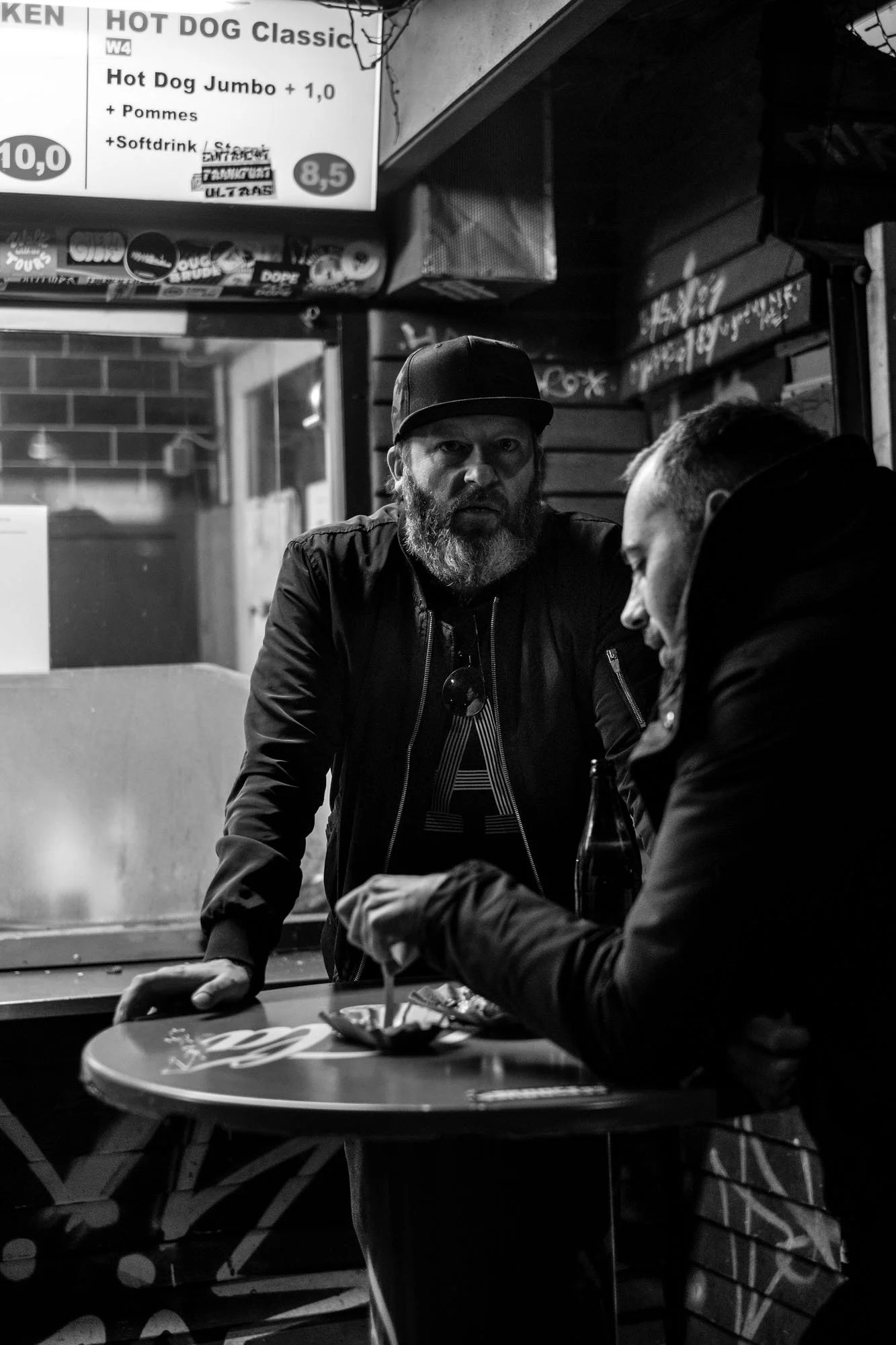  Two men talking at a standing table outside a hot dog kiosk, Berlin. Candid street portrait by Chris Perona. 