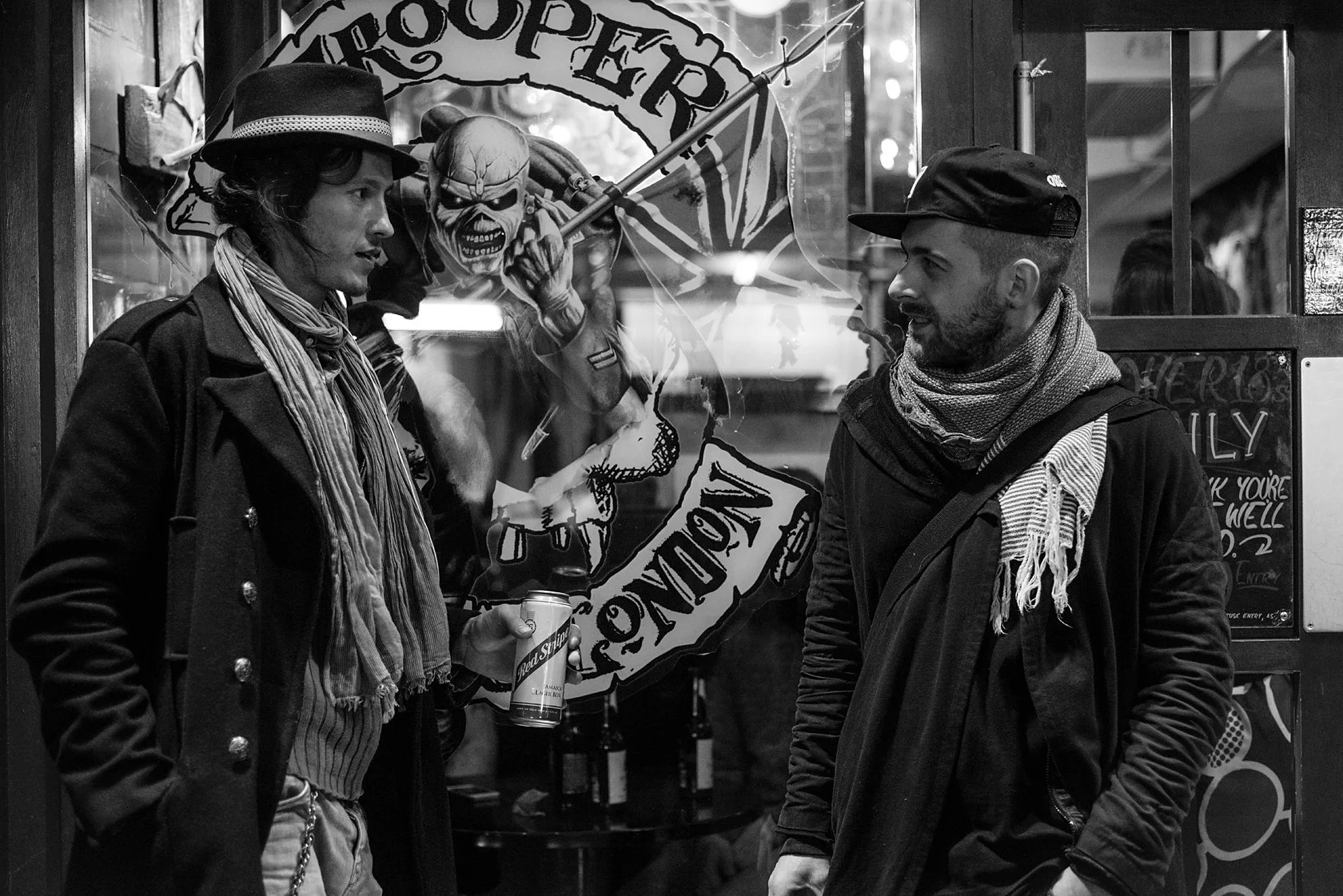  Two men in hats and scarves talking beneath a pub sign, London. Candid street portrait by Chris Perona. 