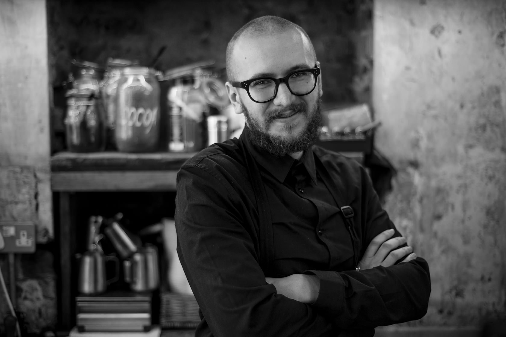  Bearded man in glasses with arms crossed in front of shelves of jars, London. Candid street portrait by Chris Perona. 