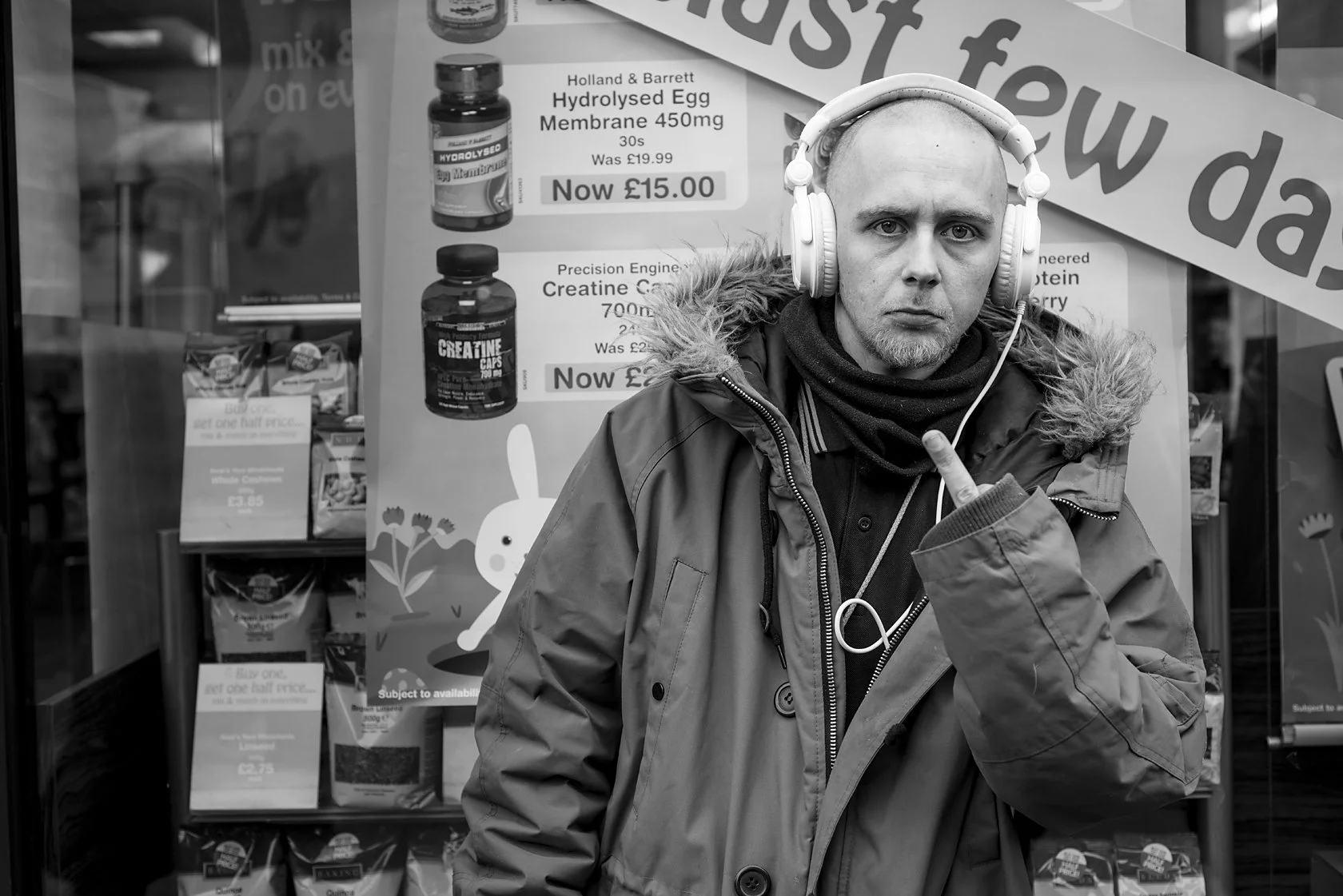  Man in a parka with headphones gesturing in front of a shop window, London. Candid street portrait by Chris Perona. 