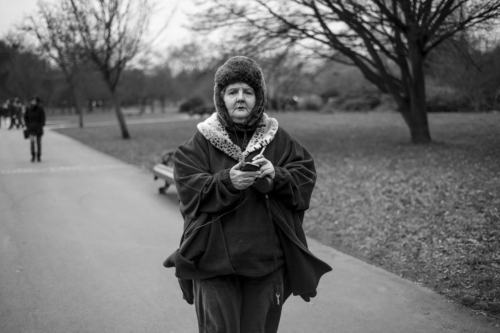  Woman in a fur-trimmed hood walking through a park, looking at her phone, London. Candid street portrait by Chris Perona. 