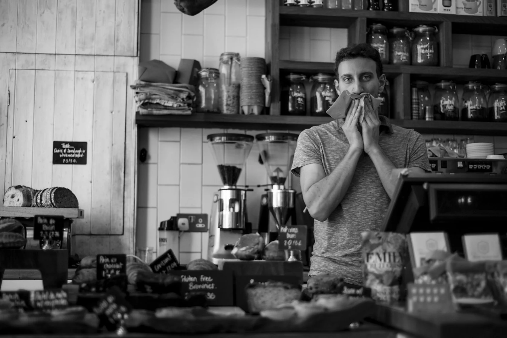  Young man laughing behind a café counter, hand to his mouth, London. Candid street portrait by Chris Perona. 