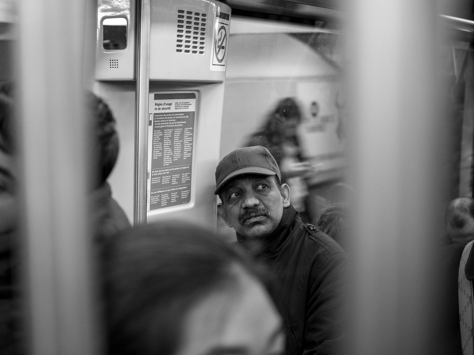  Man in a cap on a crowded metro train, seen between the poles, Paris. Candid street portrait by Chris Perona. 