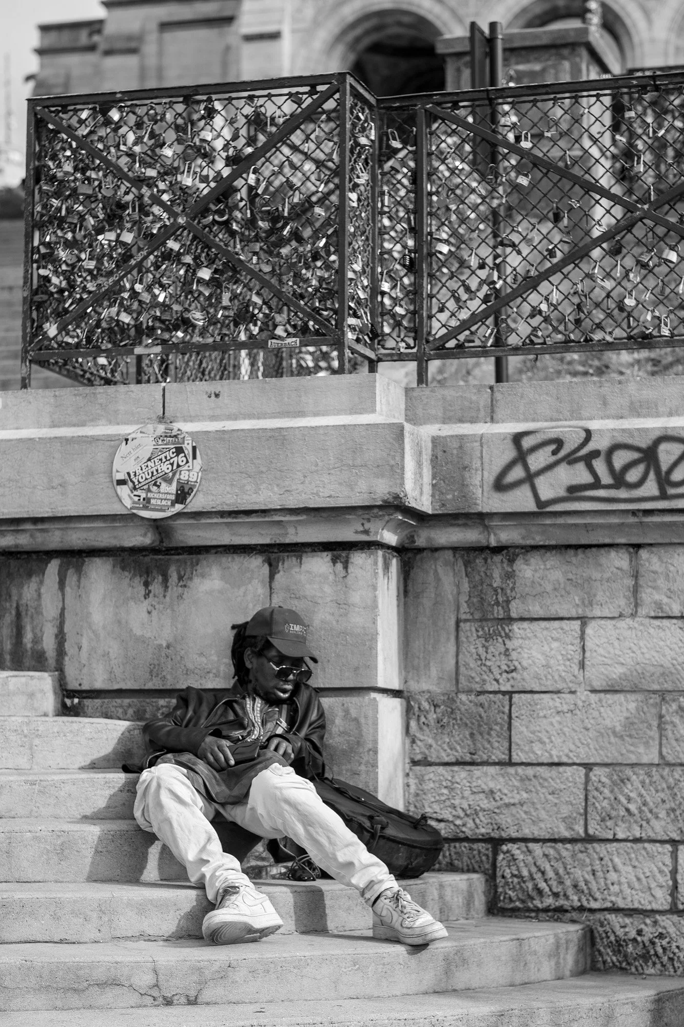  Man in a cap sitting on stone steps beneath a railing covered in love padlocks, Paris. Candid street portrait by Chris Perona. 