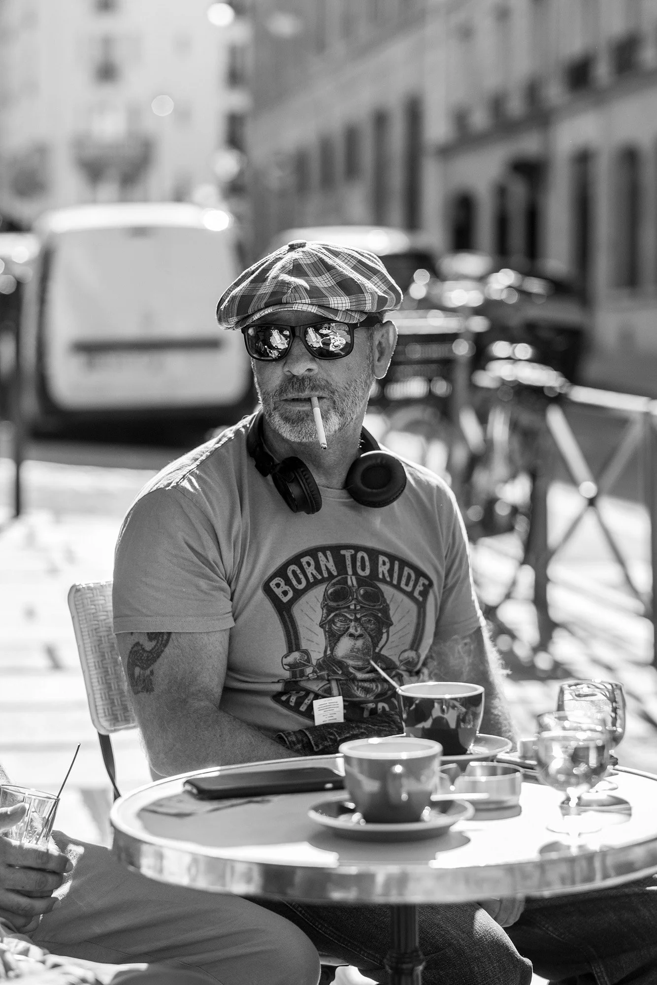  Man in a cap and mirrored sunglasses smoking at a café table, Paris. Candid street portrait by Chris Perona. 