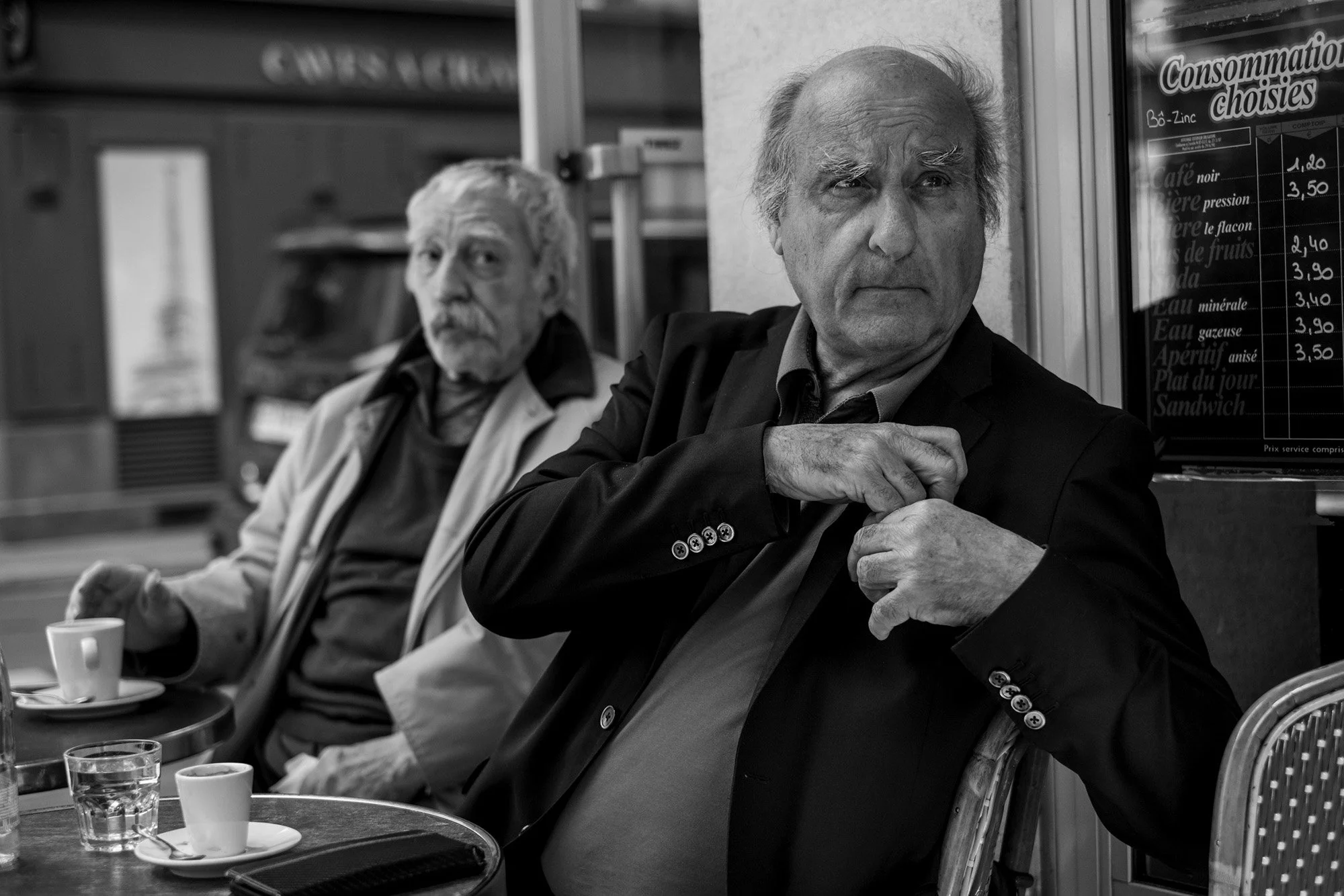  Two older men at a café terrace, one adjusting his shirt, Paris. Candid street portrait by Chris Perona. 