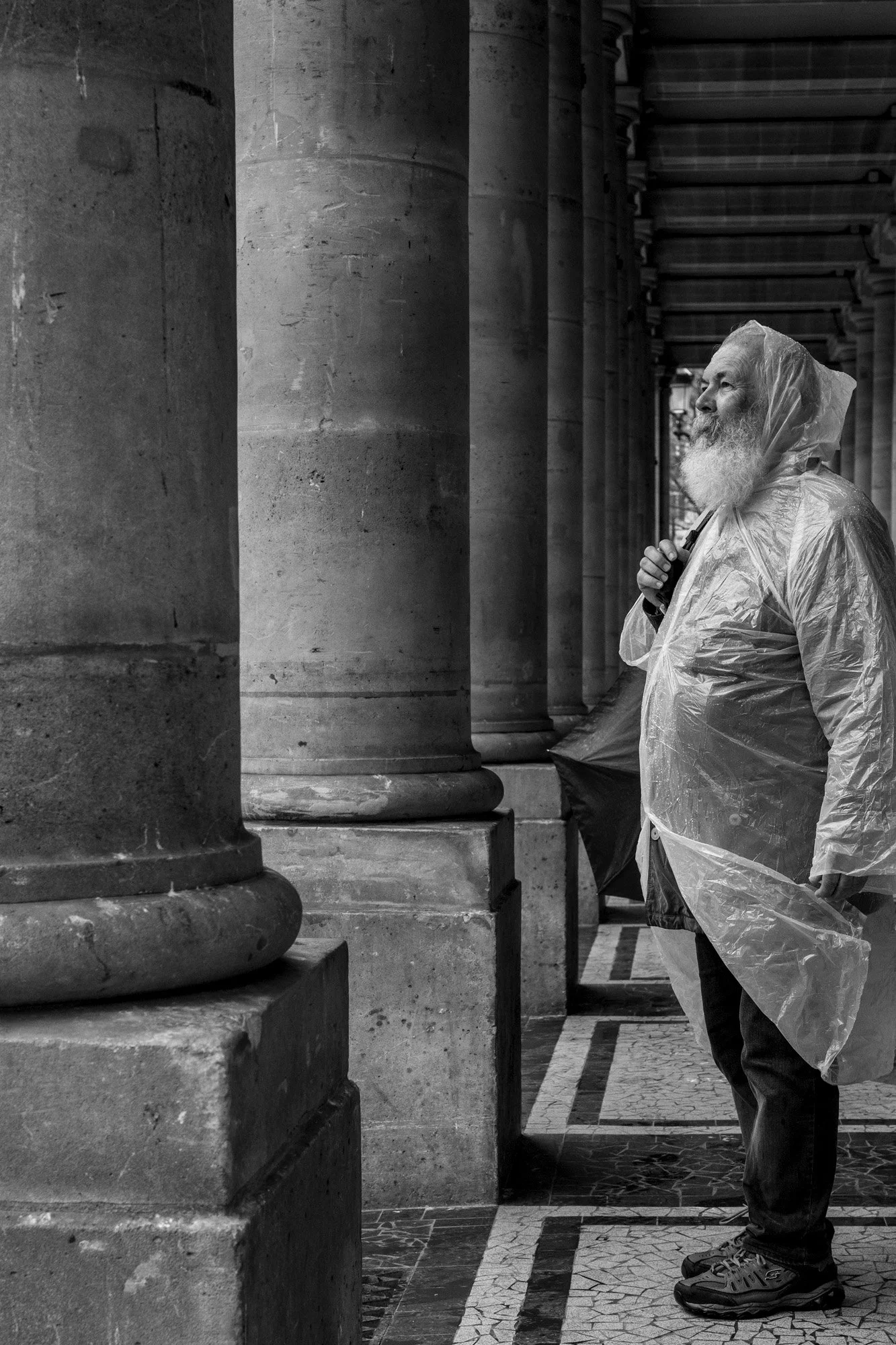  Older man with a long white beard in a rain poncho beneath a stone colonnade, Paris. Candid street portrait by Chris Perona. 