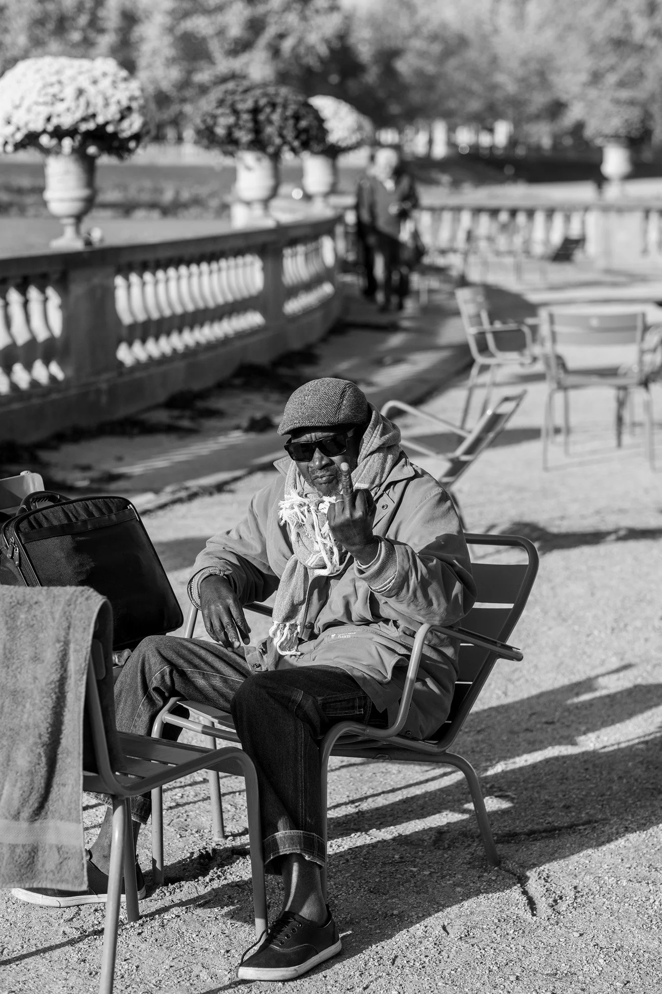  Man in a cap and sunglasses relaxing on a metal chair in a Paris garden. Candid street portrait by Chris Perona. 