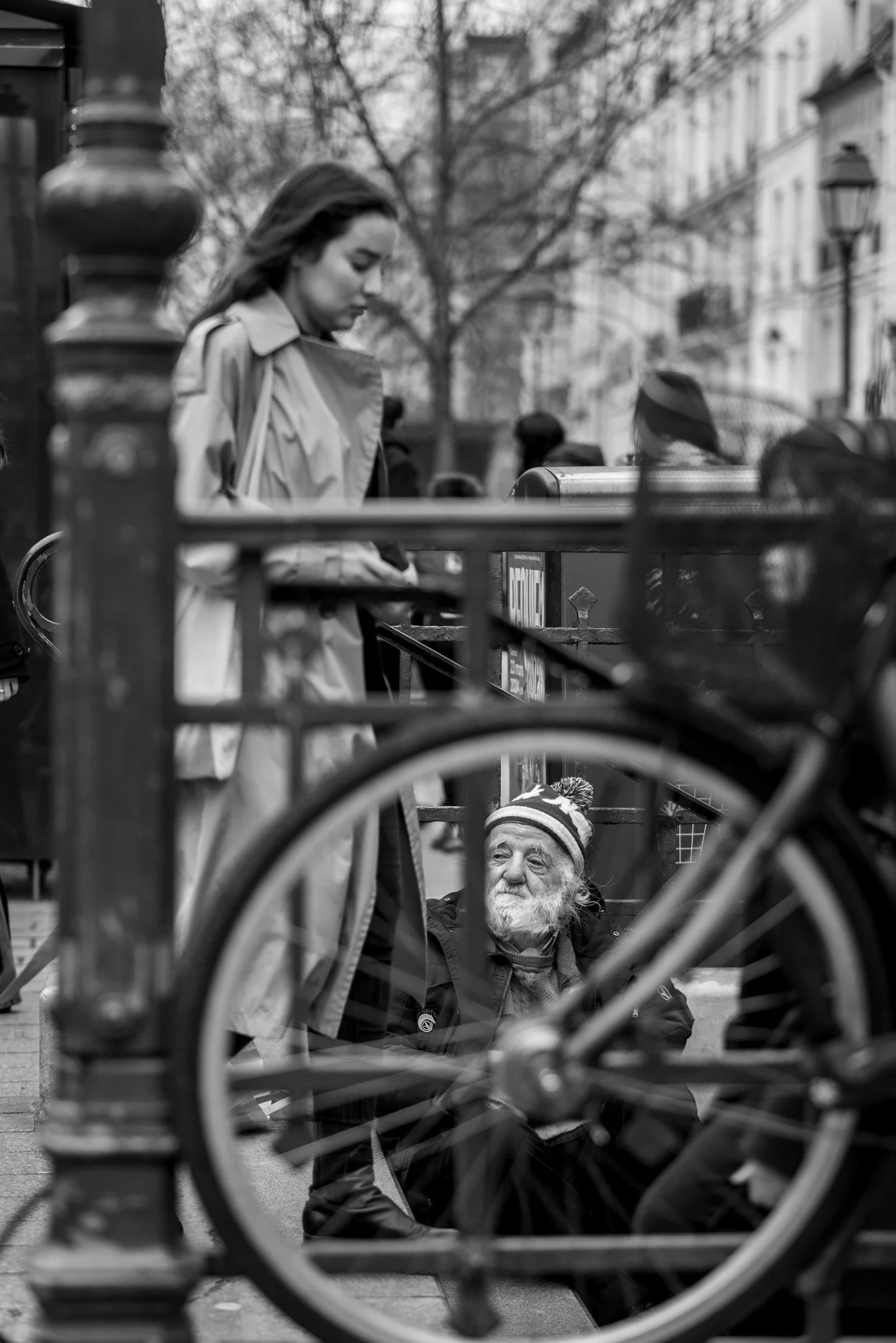  Older man in a striped beanie sitting by a metro railing as a woman walks past, Paris. Candid street portrait by Chris Perona. 