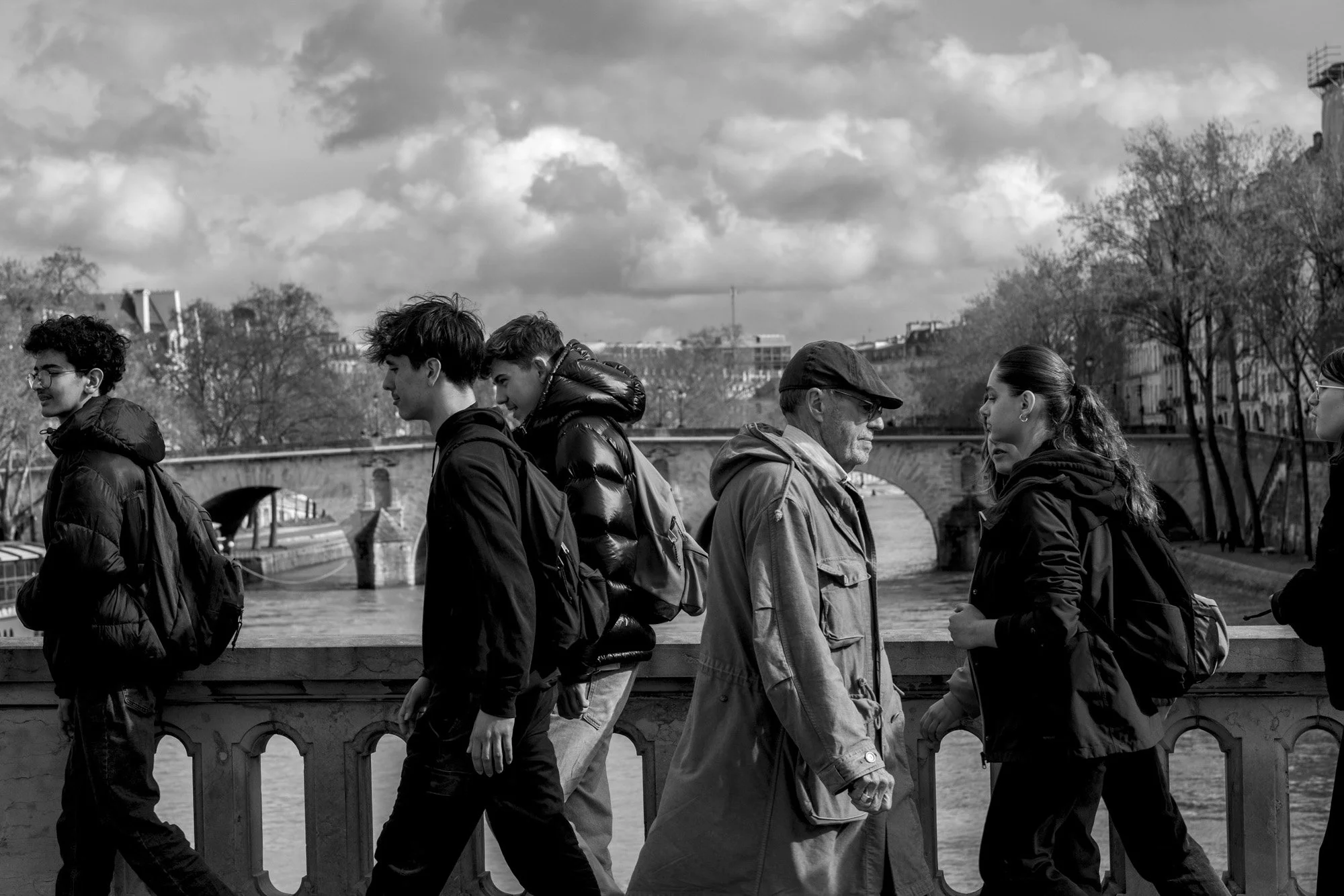  Passers-by crossing a bridge over the Seine under a cloudy sky, Paris. Street photograph by Chris Perona. 
