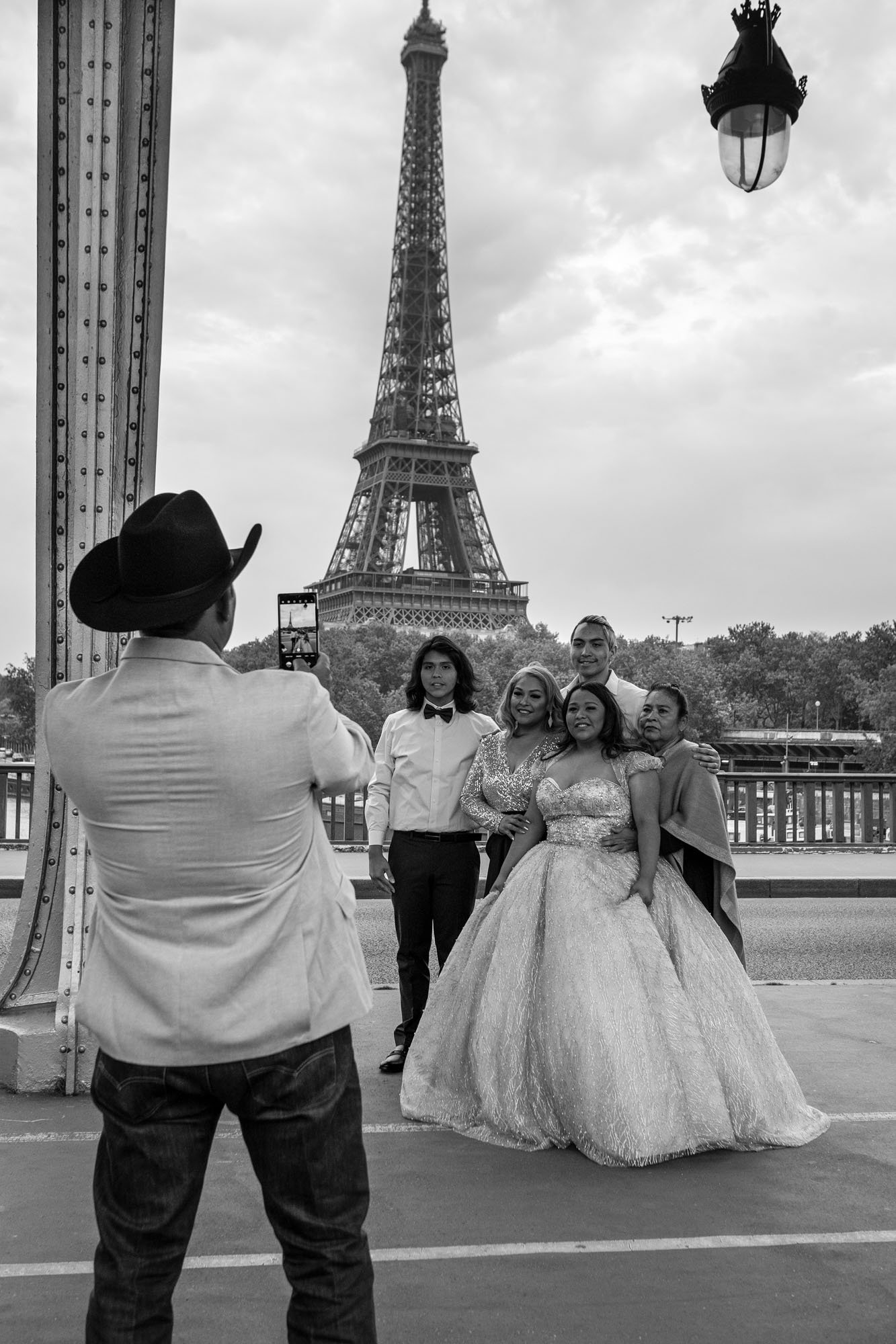  Man in a cowboy hat photographing a family and a girl in a ball gown before the Eiffel Tower, Pont de Bir-Hakeim, Paris. Street photograph by Chris Perona. 