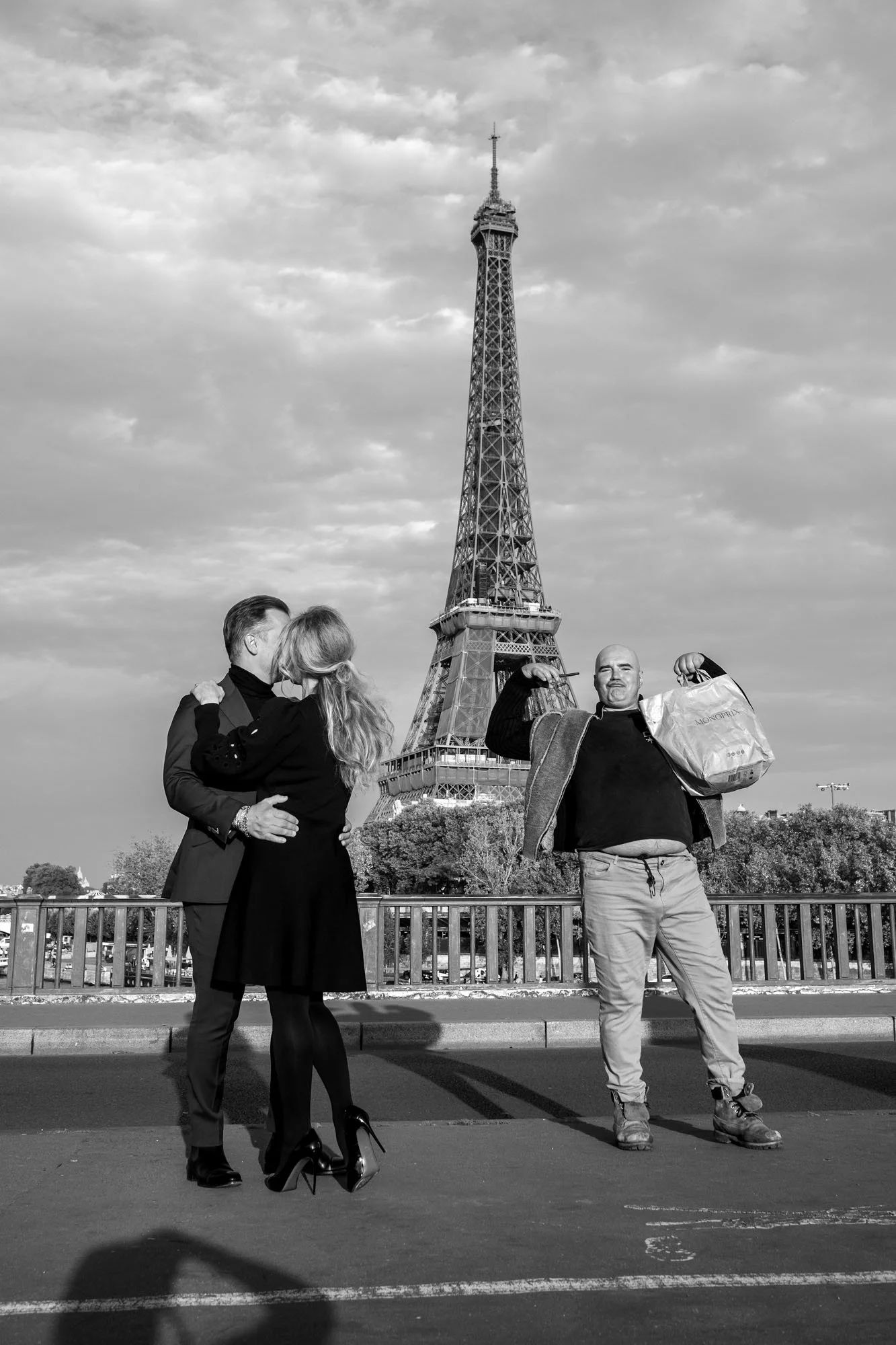  Couple kissing beside a man posing with open arms before the Eiffel Tower, Pont de Bir-Hakeim, Paris. Street photograph by Chris Perona. 