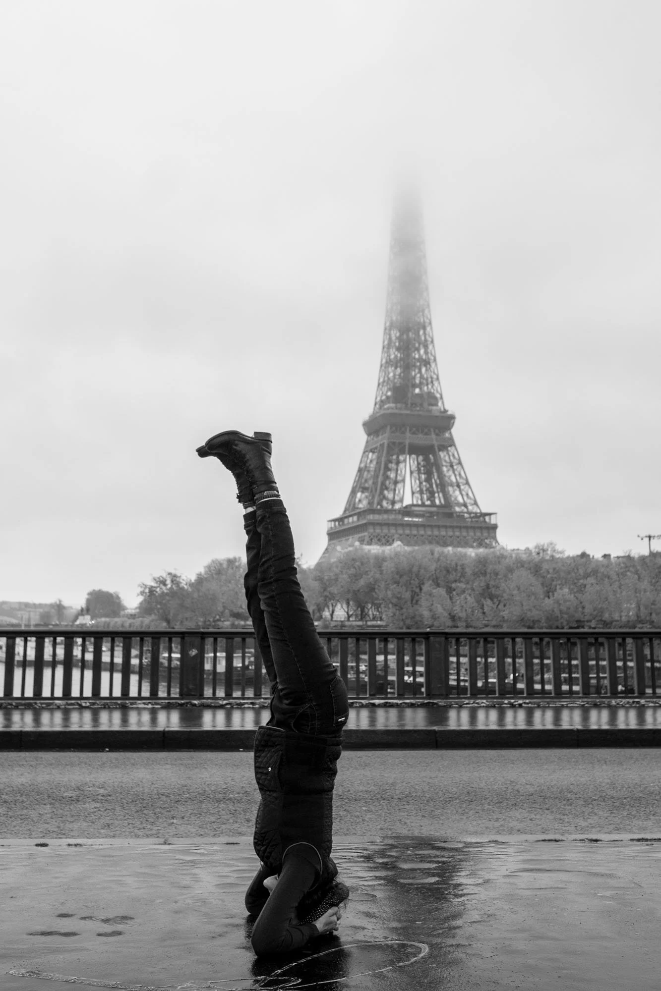  Person doing a headstand on the wet pavement, the Eiffel Tower lost in fog, Pont de Bir-Hakeim, Paris. Street photograph by Chris Perona. 