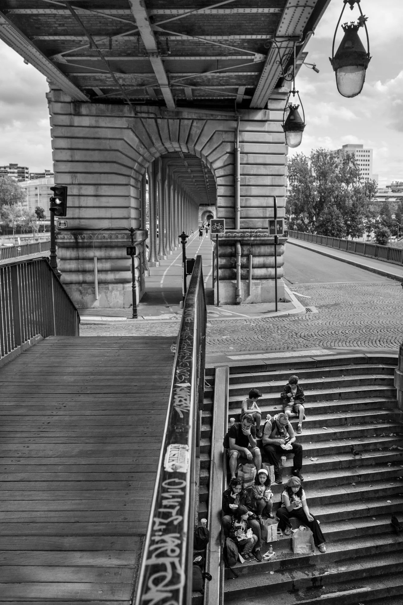  People sitting on the stairs beneath the stone pier and the metro deck, Pont de Bir-Hakeim, Paris. Street photograph by Chris Perona. 