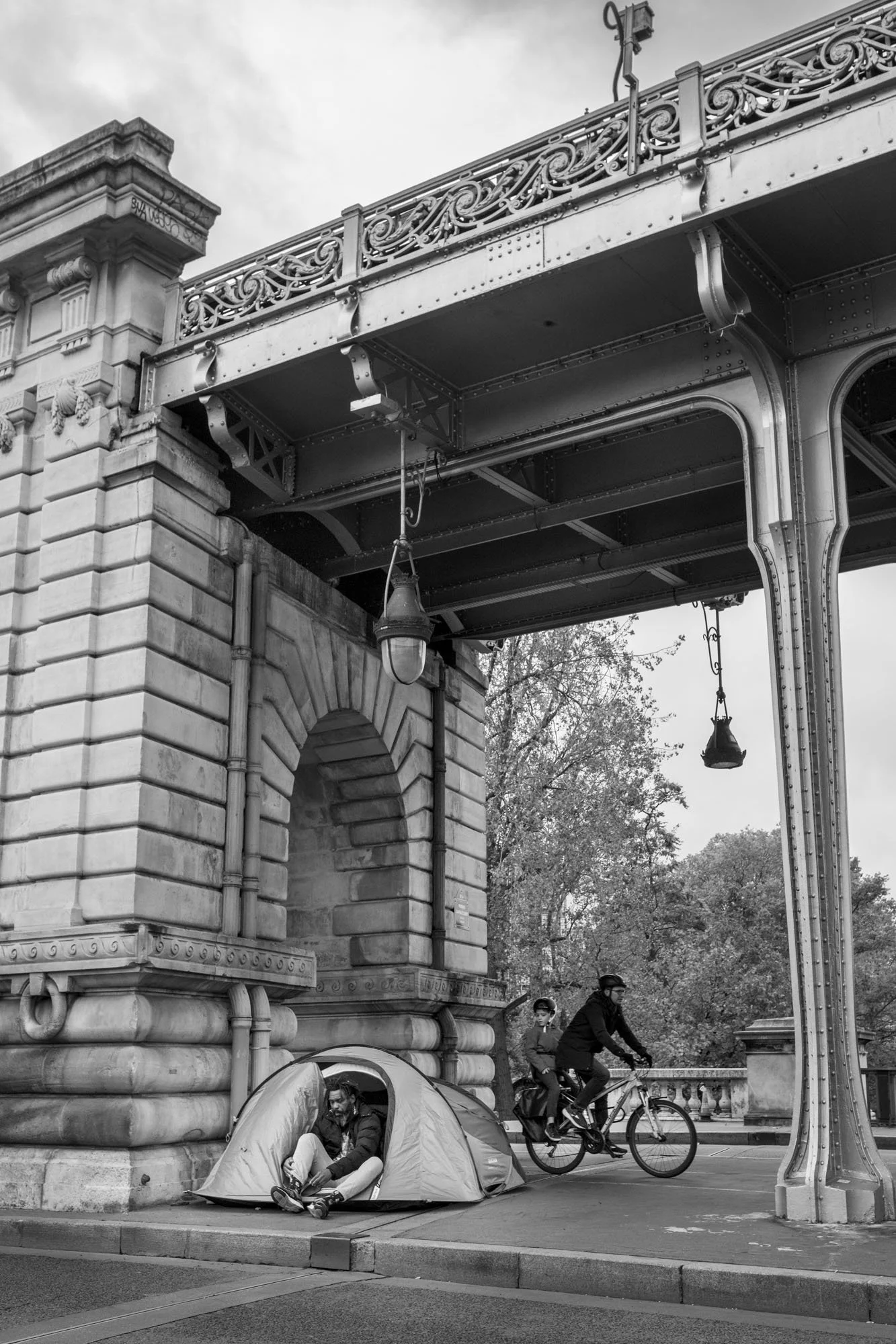  A tent pitched beneath the stone pier as a cyclist rides past, Pont de Bir-Hakeim, Paris. Street photograph by Chris Perona. 