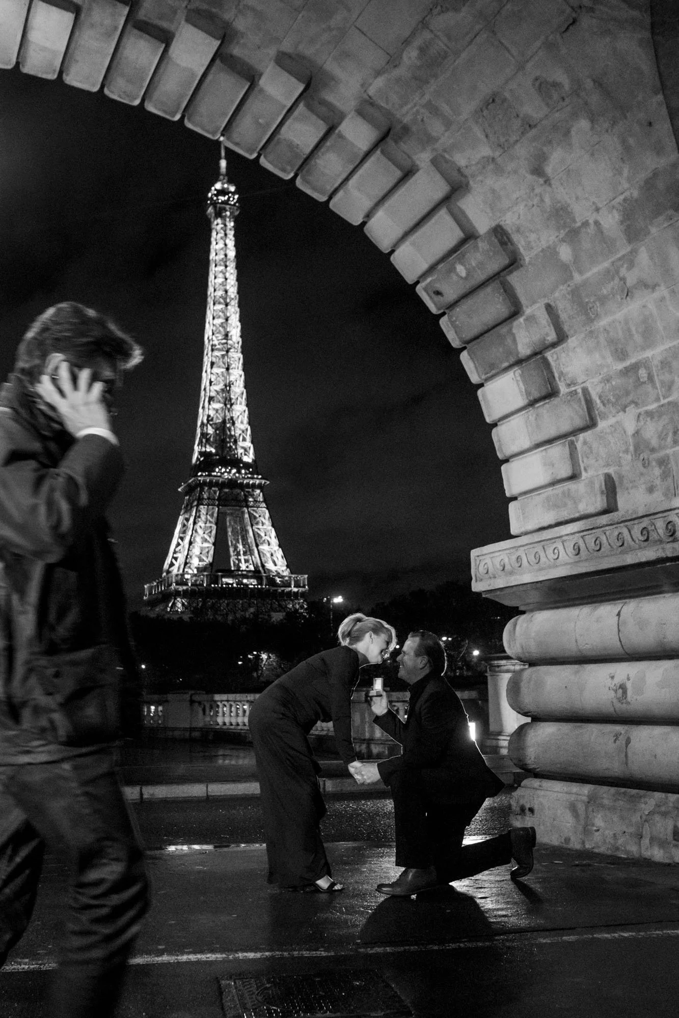  Marriage proposal at night beneath a stone arch, the Eiffel Tower lit up, Pont de Bir-Hakeim, Paris. Street photograph by Chris Perona. 