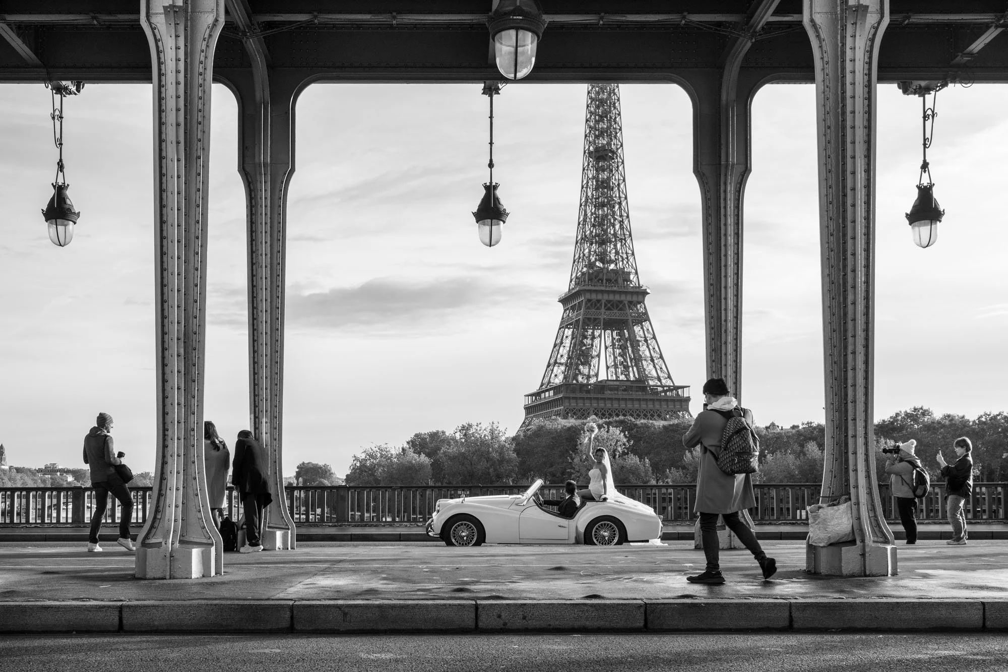  Vintage convertible passing between iron columns that frame the Eiffel Tower, Pont de Bir-Hakeim, Paris. Street photograph by Chris Perona. 