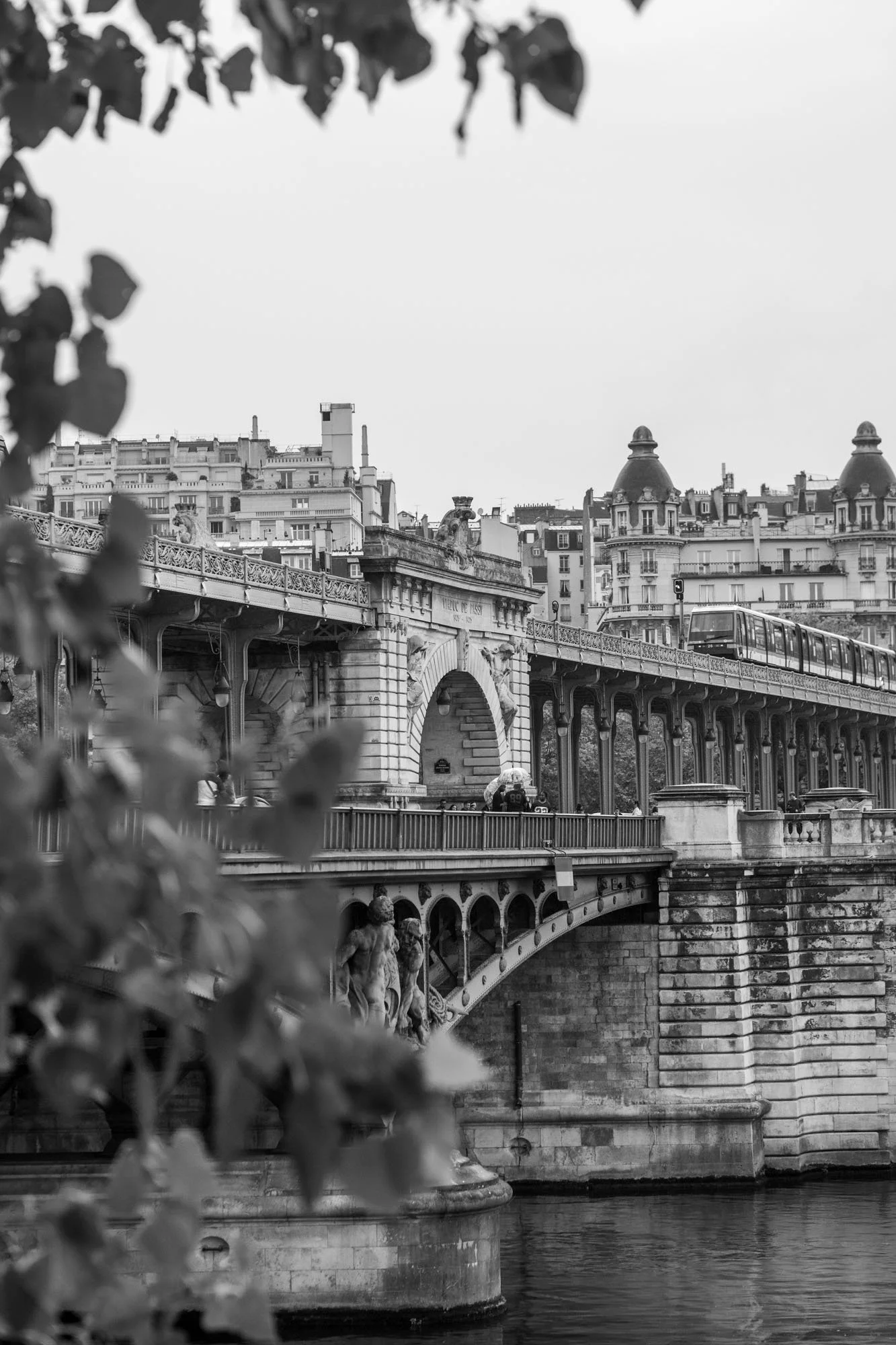  Stone arches of the Pont de Bir-Hakeim framed by leaves, with the buildings of Passy, Paris. Architecture photograph by Chris Perona. 