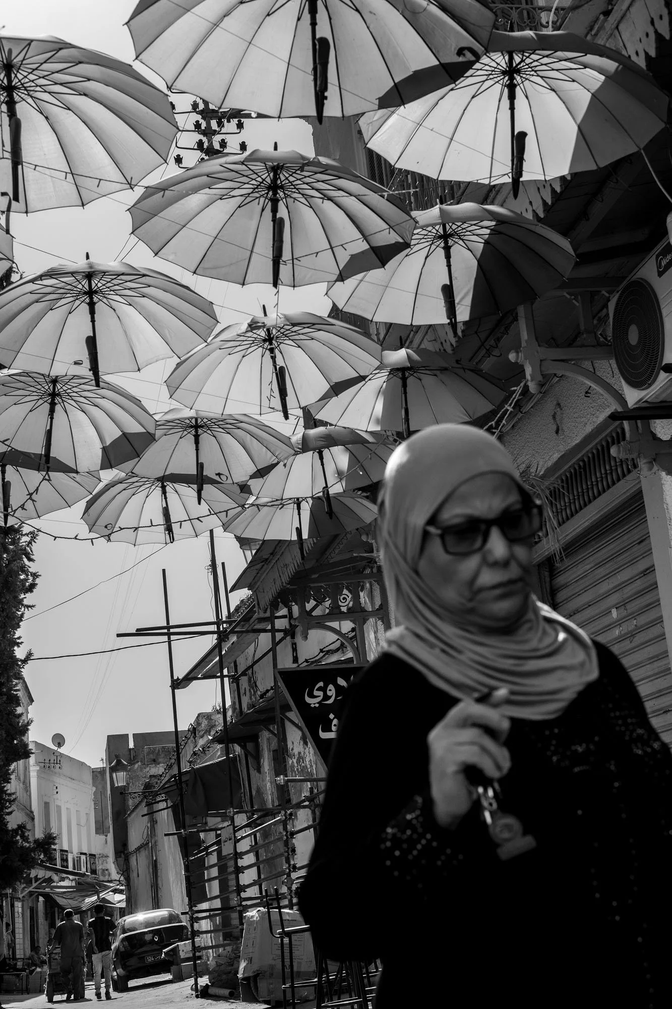  Woman in a headscarf and sunglasses beneath an alley of suspended umbrellas, medina of Tunis. Black-and-white street photograph by Chris Perona. 
