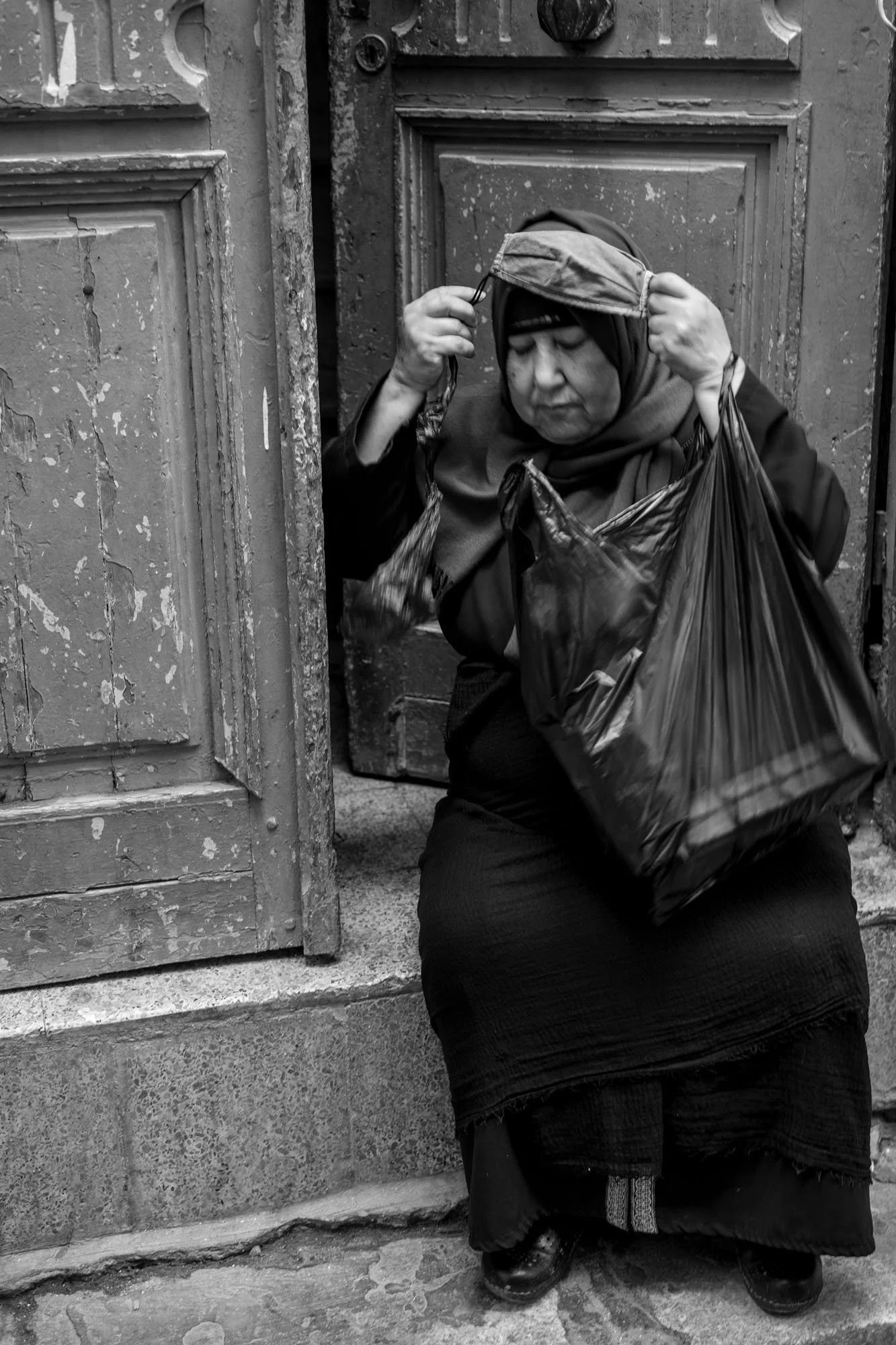  Older woman adjusting her headscarf, seated beside an old wooden door, medina of Tunis. Black-and-white street photograph by Chris Perona. 