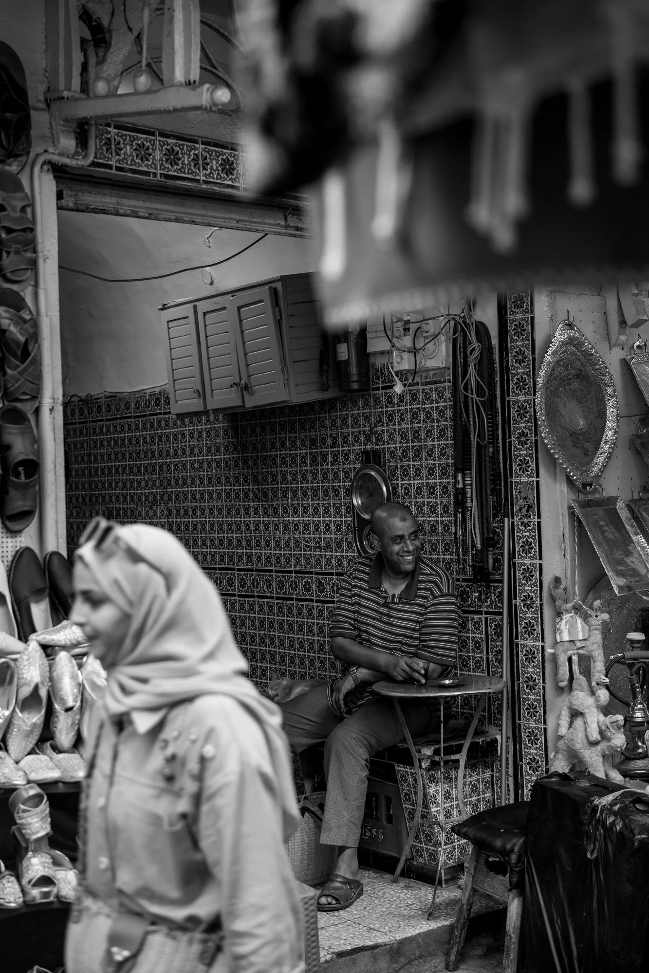  Woman in a headscarf walking past a tiled shop and its seated shopkeeper, medina of Tunis. Black-and-white street photograph by Chris Perona. 