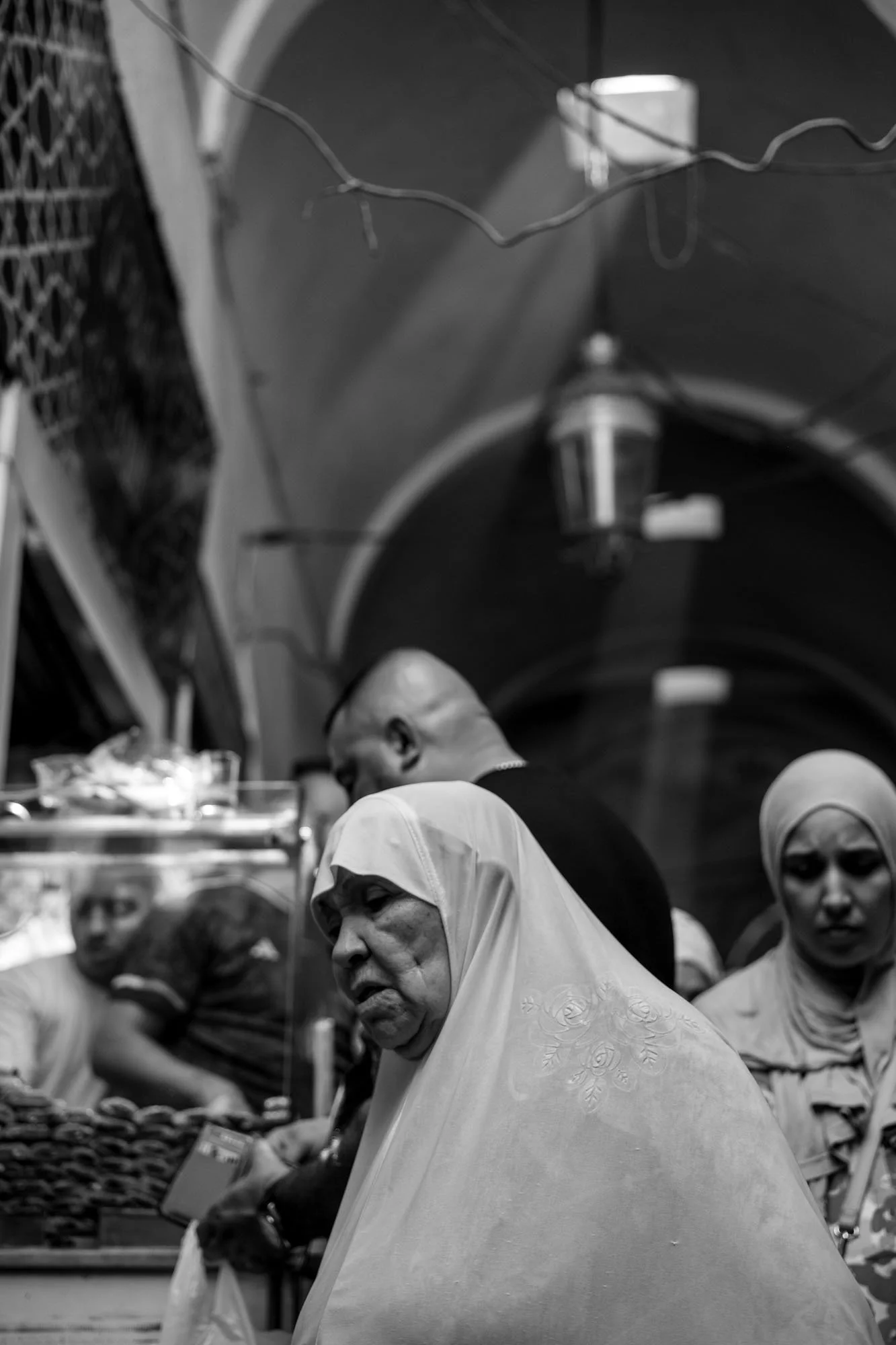  Older woman in a white veil among the crowd of a vaulted souk, medina of Tunis. Black-and-white street photograph by Chris Perona. 